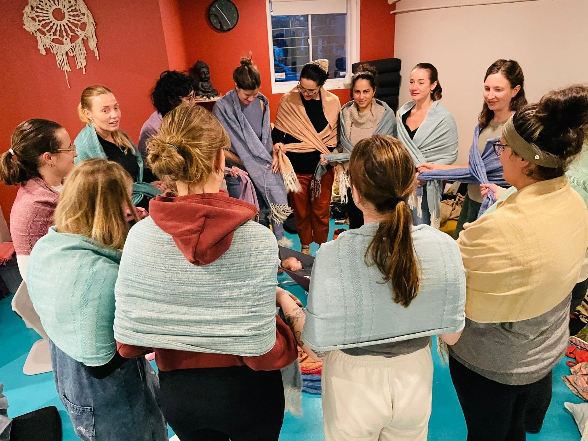 A group of women standing in a circle holding scarves in a cozy, warmly lit room that has a window, a wall clock, and a decorative wall hanging.
