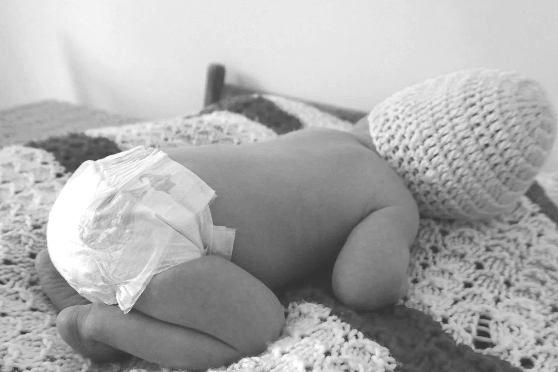 A baby lying on a bed in a diaper and knit hat, resting on a crocheted blanket.
