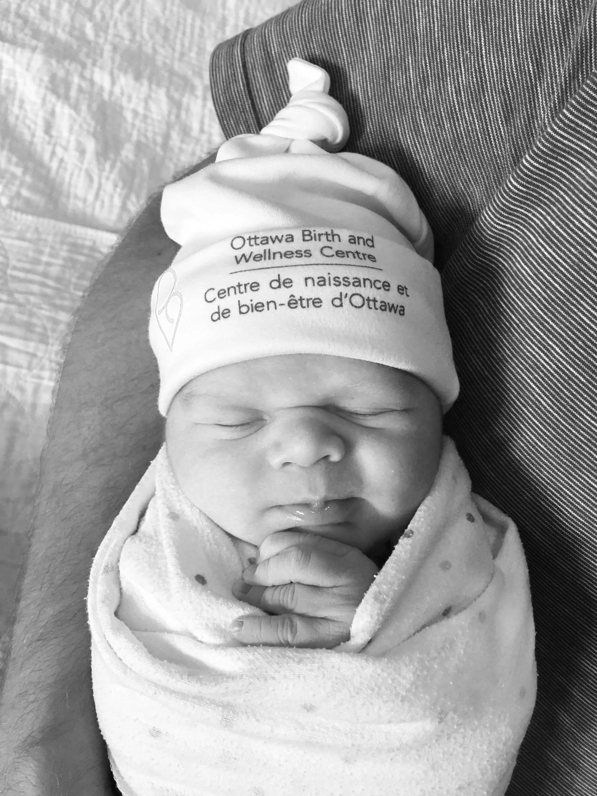 Black and white photo of a newborn baby wrapped in a blanket, wearing a hat that reads Ottawa Birth and Wellness Centre in both English and French, sleeping peacefully.