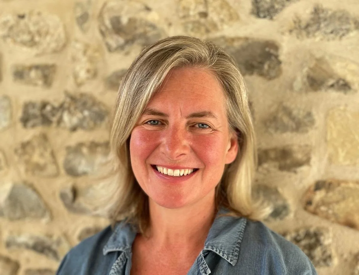 A smiling woman with shoulder-length blonde hair standing in front of a stone wall.