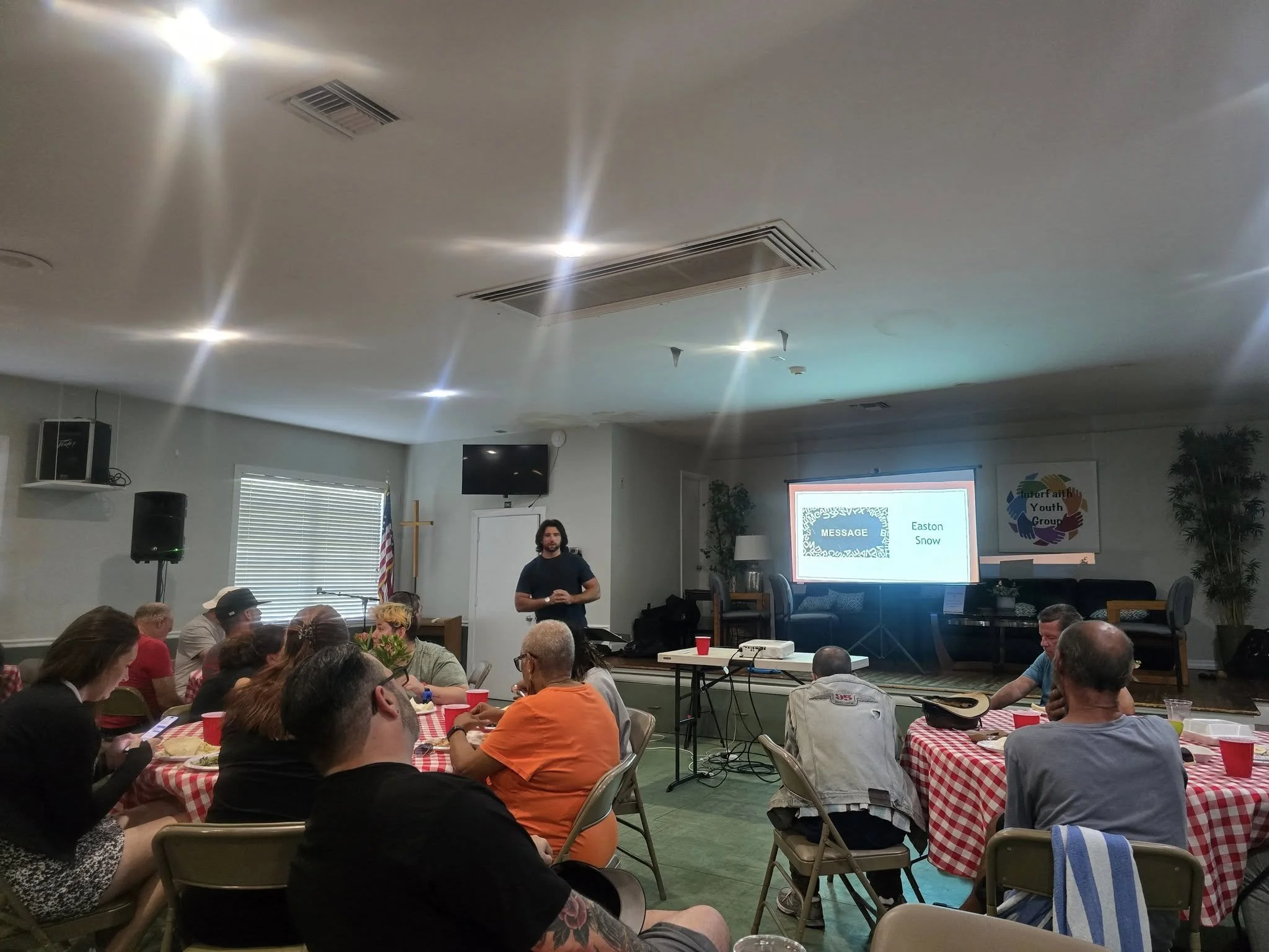 A group of people sitting at tables with red and white checkered tablecloths in a meeting room, listening to a man standing in front of a screen displaying a presentation titled 'MESSAGE' with the name 'Easton Snow' on it. There are plants, a cross, and an American flag in the background.