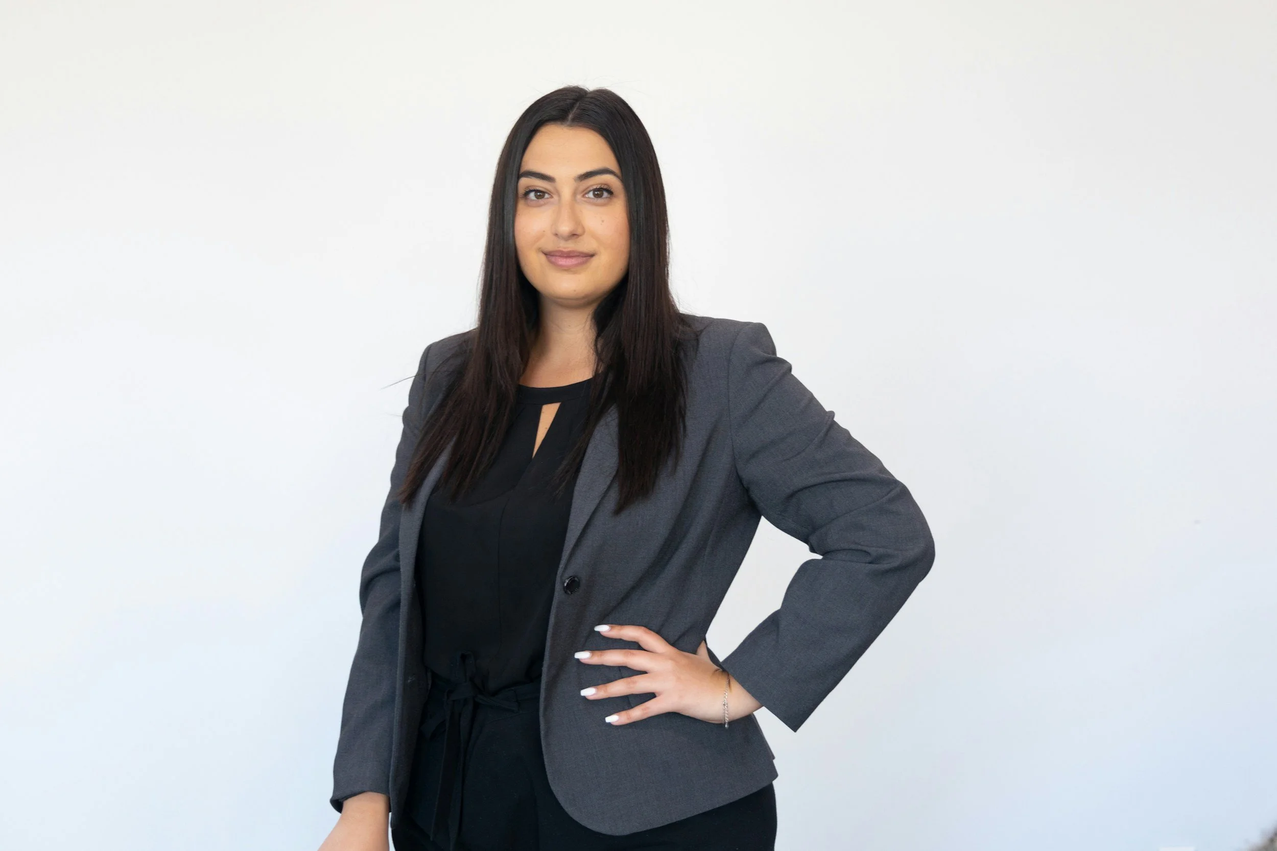 A woman with long dark hair wearing a dark blouse and gray blazer, standing against a plain white background with one hand on her hip.