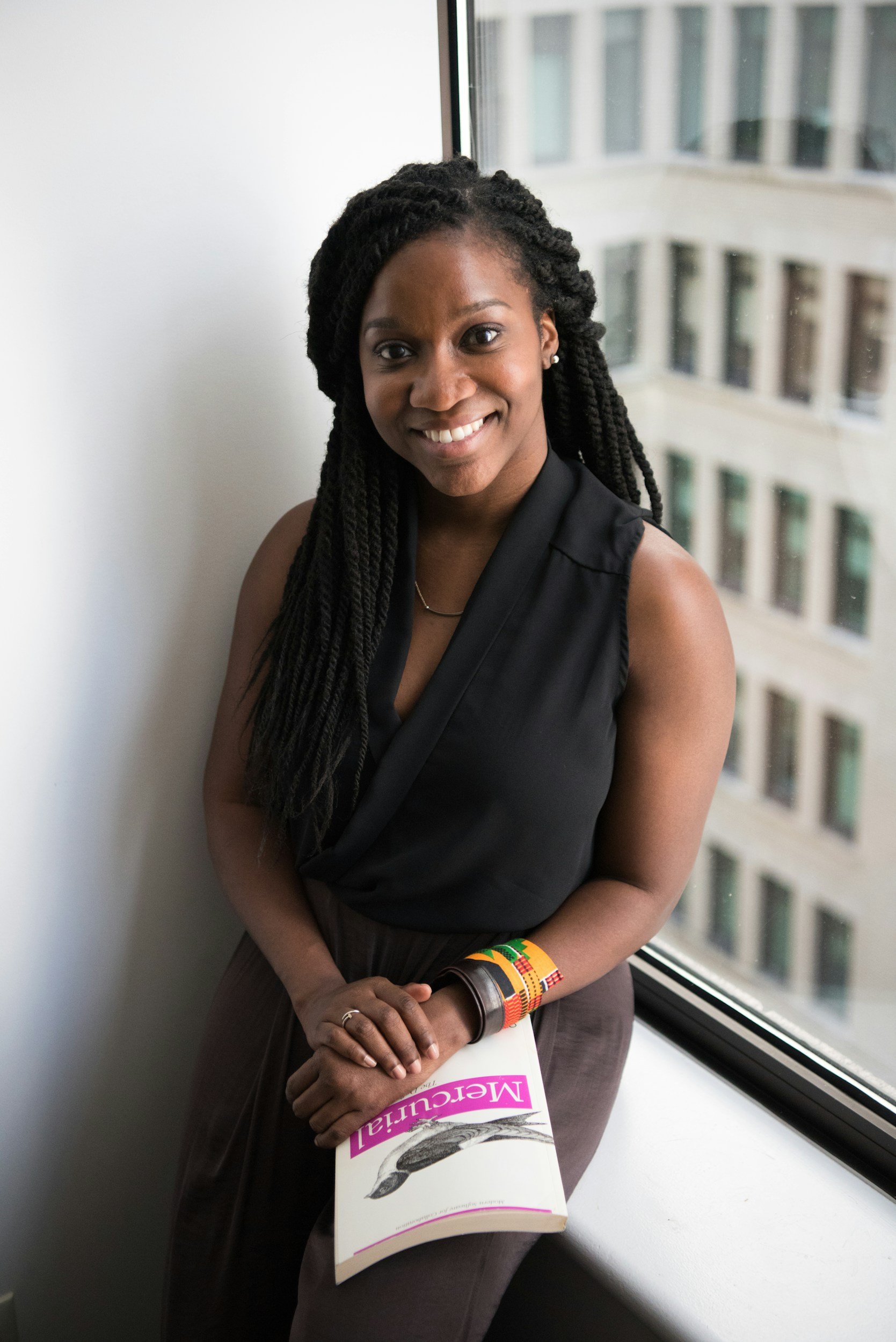 A woman with dark braided hair and a black sleeveless top standing near a window, holding a brochure titled "Mercantil".
