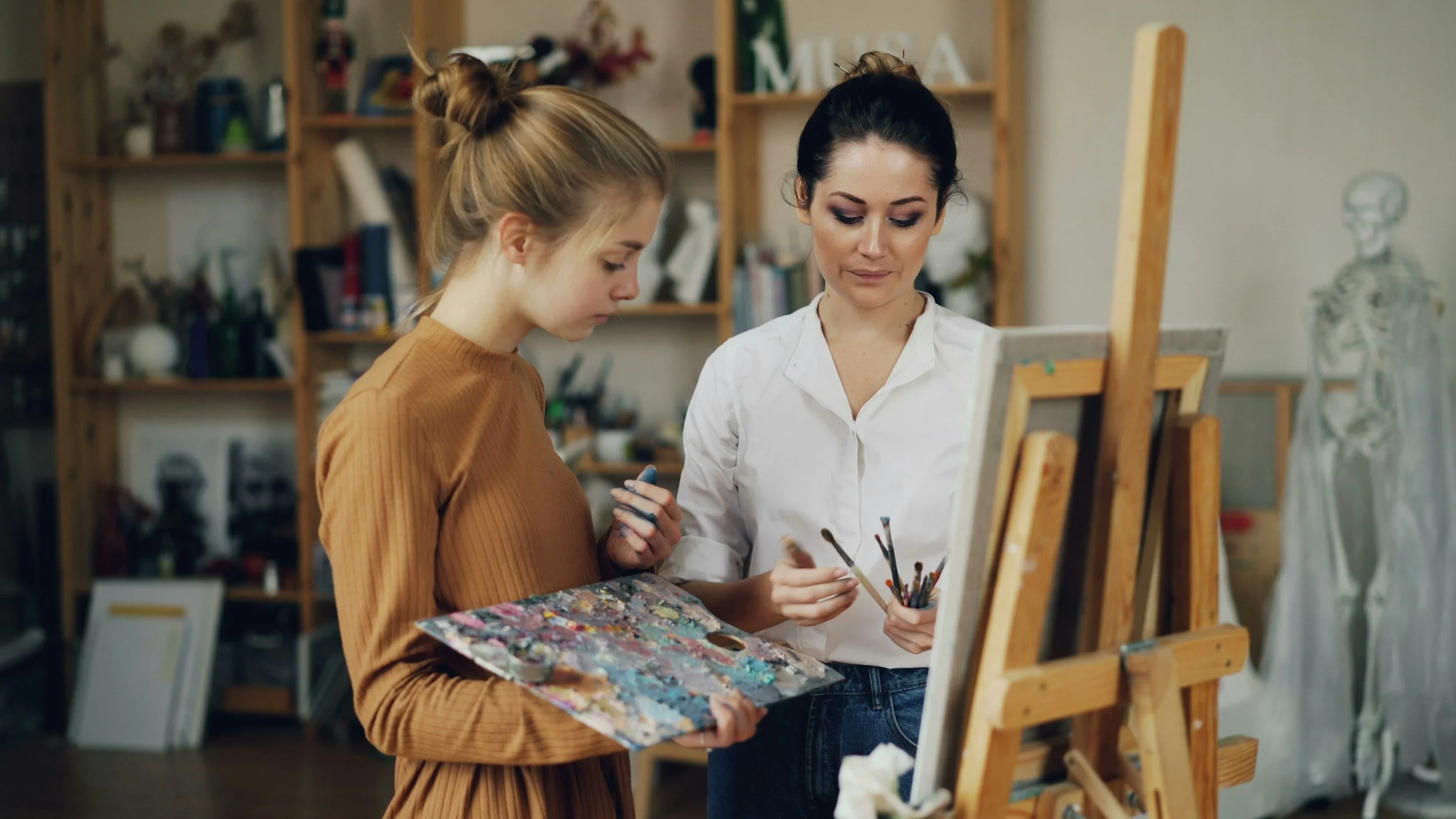 Two women in an art studio discussing a painting. One woman in a brown shirt holds a palette with paint, while the other woman in a white shirt holds paintbrushes. Art supplies and a skeleton model are visible in the background.