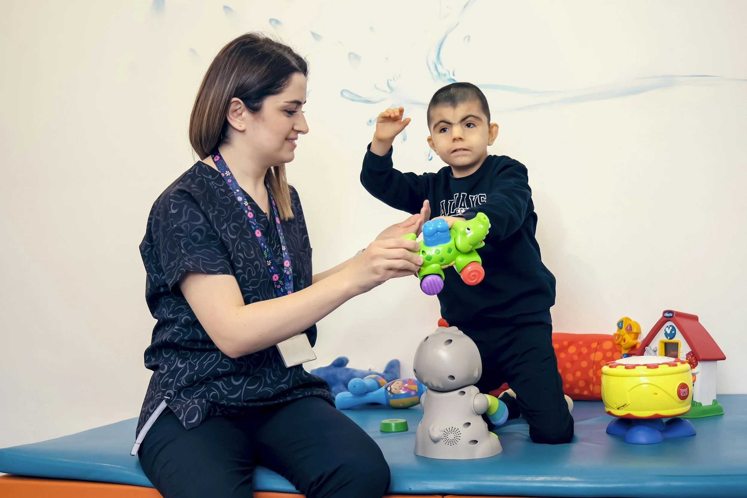 A woman and a young boy sitting on a blue mat playing with colorful toys. The woman has brown hair and is smiling, while the boy has short dark hair and is making a facial expression. There are various toys on the mat, including a robot, a yellow toy with a red lid, and other play items.