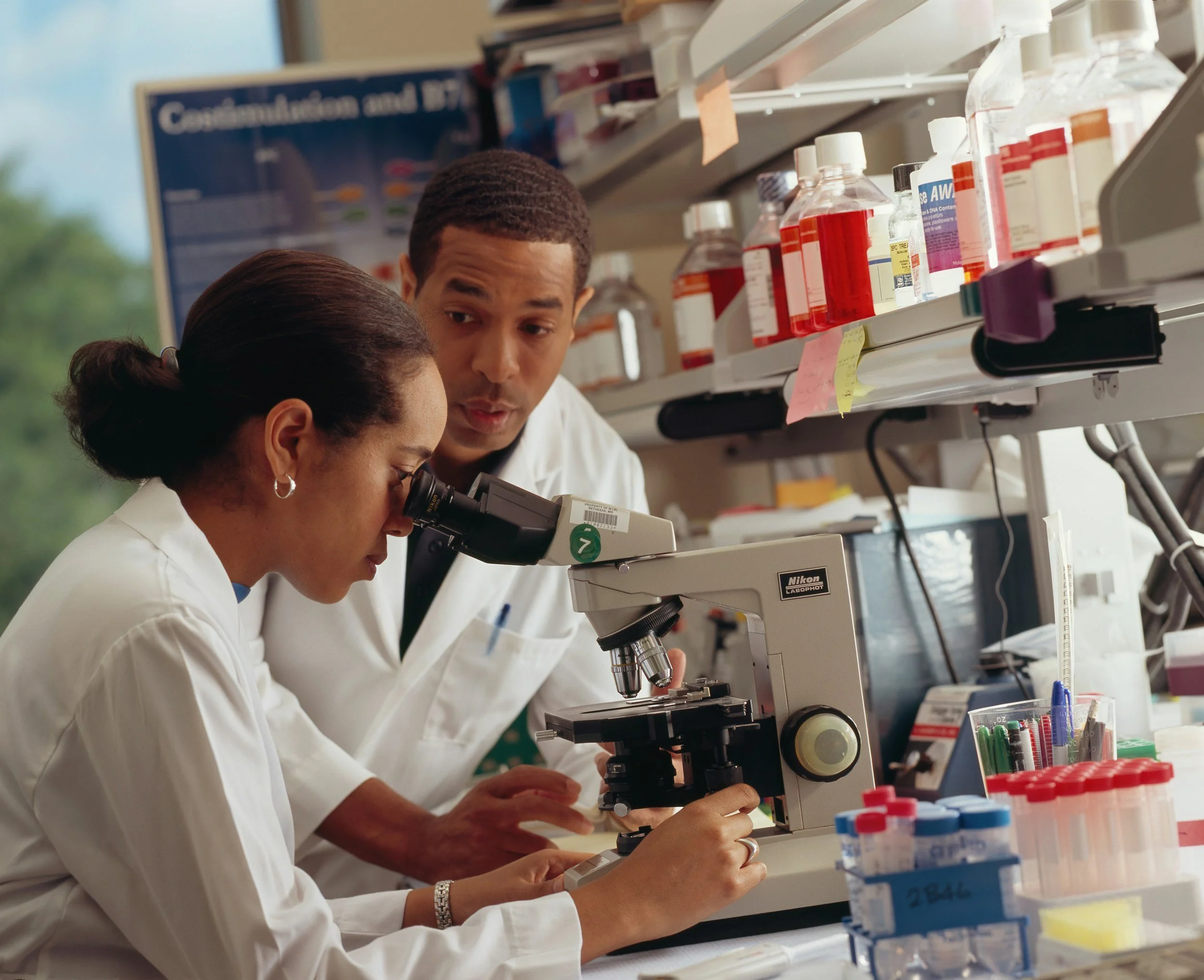 Two scientists in white lab coats working with a microscope in a laboratory filled with chemical bottles and lab equipment.