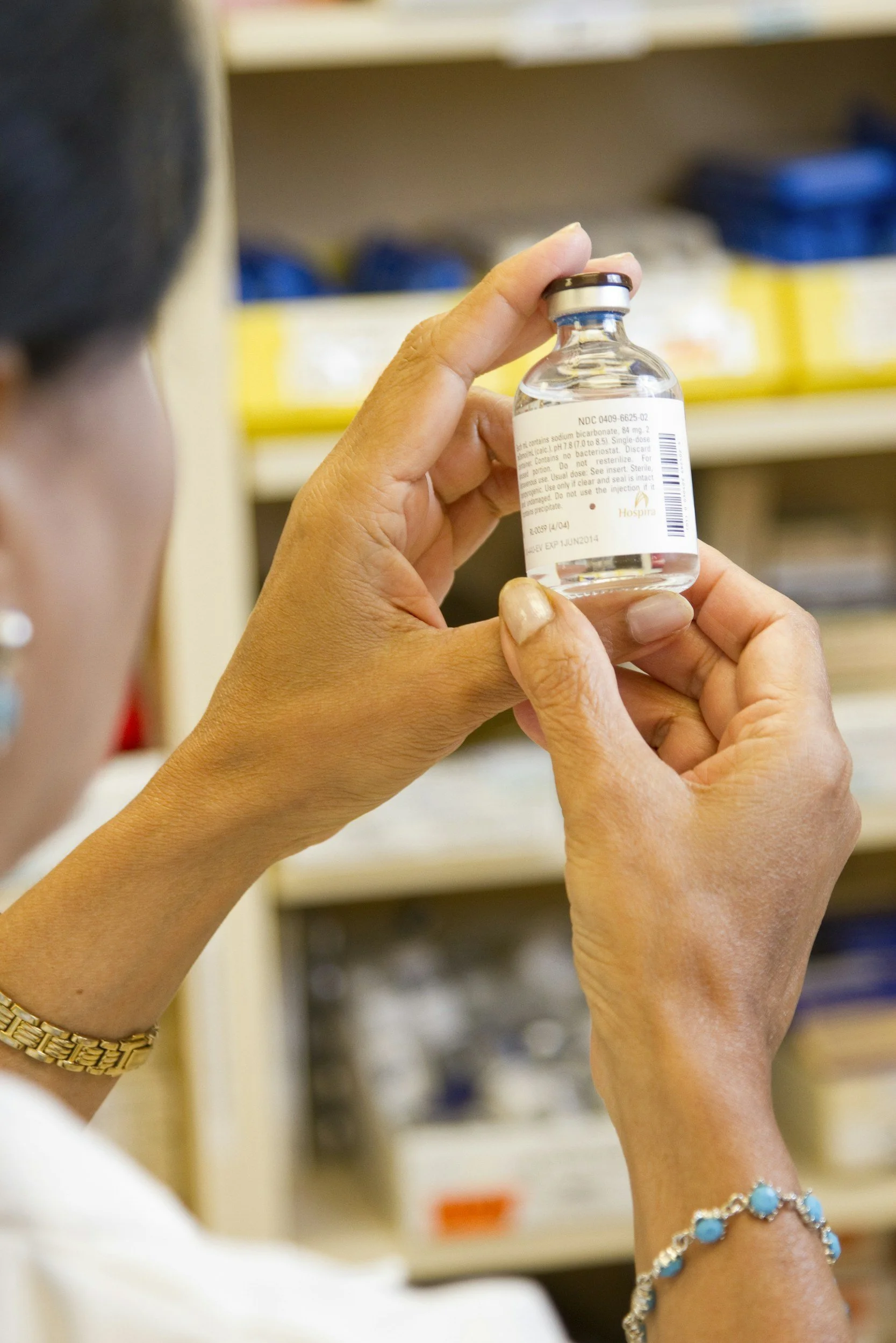 A person holding a small vial of clear liquid with a label, in a pharmacy or medical setting.