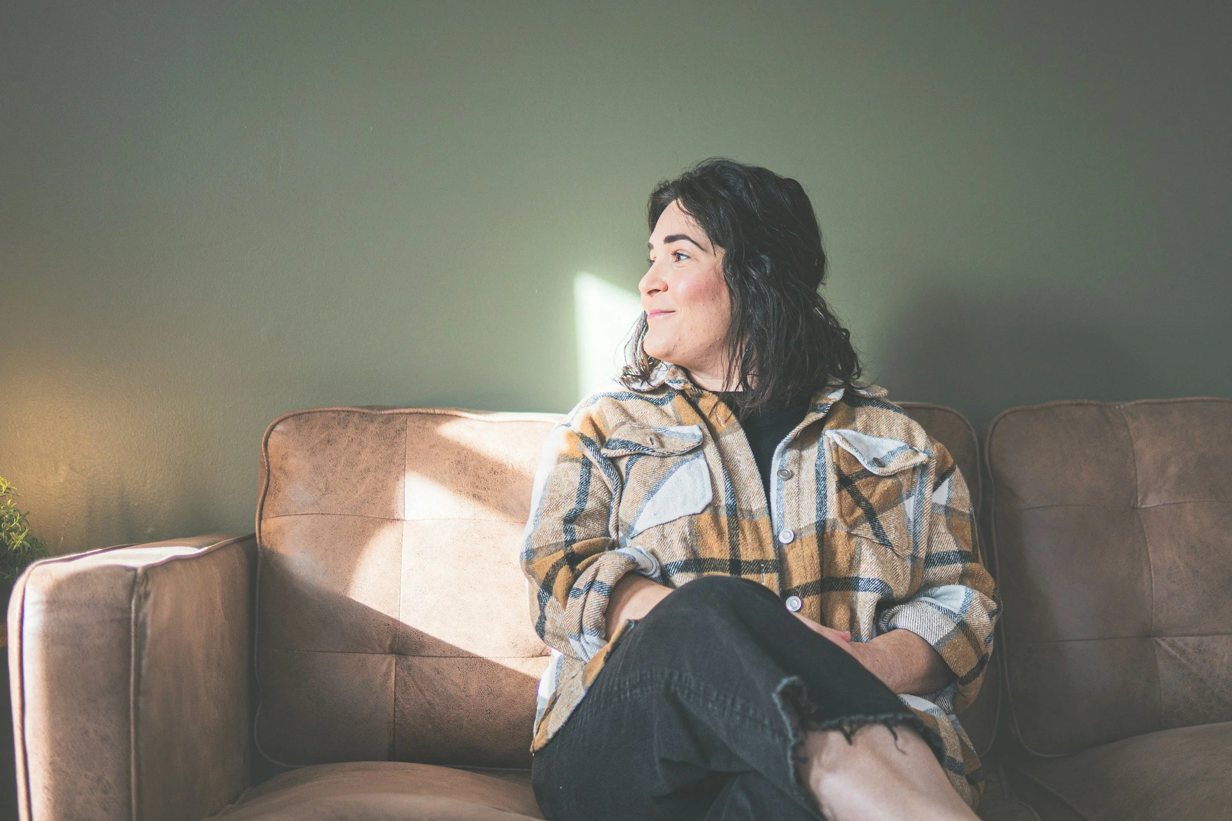 A woman with dark wavy hair, wearing a checkered jacket, is sitting on a brown sofa with her legs crossed. She is looking towards the right, with sunlight illuminating part of her face. The background features a plain green wall, and there is a small plant visible at the bottom left corner.