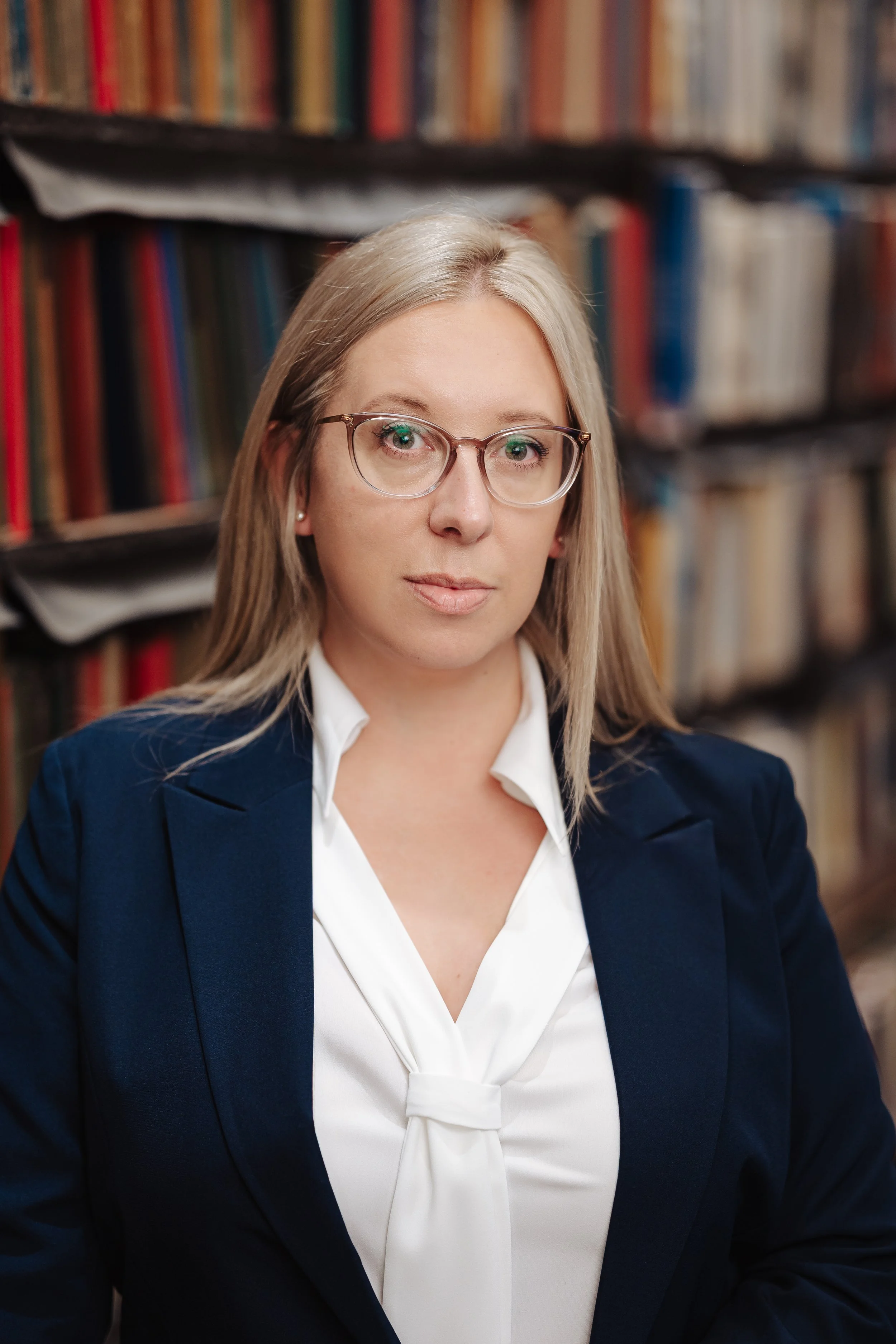 A woman with blonde hair wearing glasses, a white blouse with a bow, and a dark blazer, sitting in front of a bookshelf filled with books.