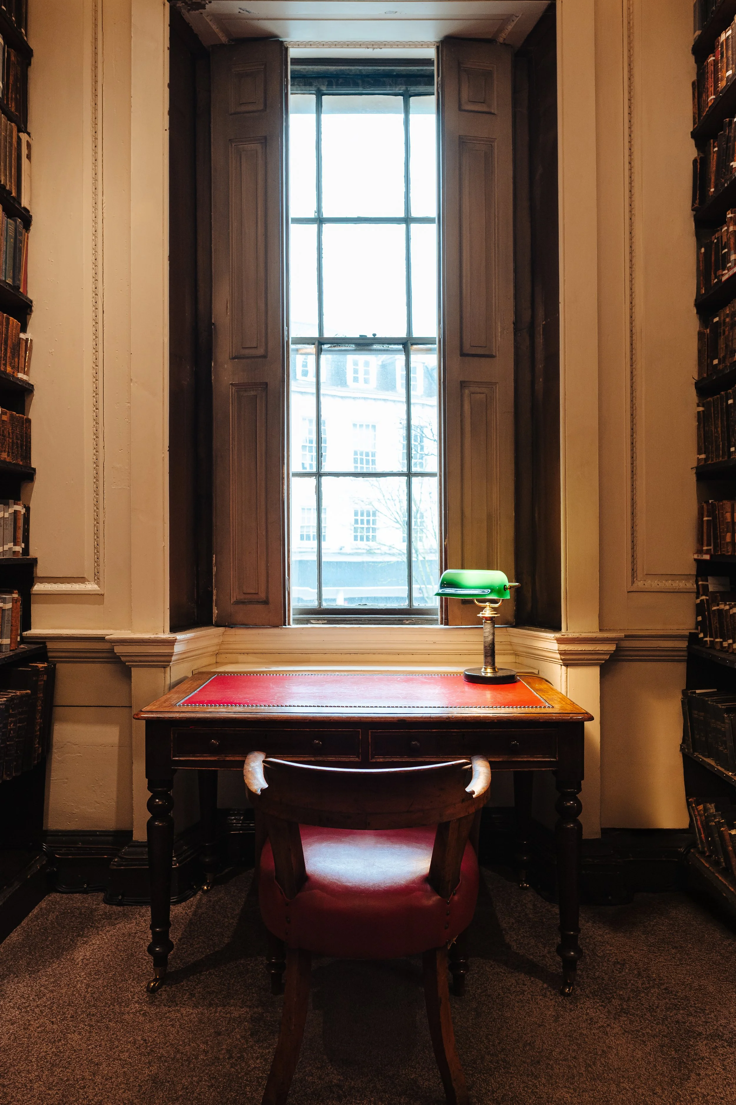 A study room with a window behind a wooden desk and a red leather armchair. The desk has a green banker’s lamp and is flanked by tall bookshelves filled with books. The room has ornate wall moldings and dark carpeting.