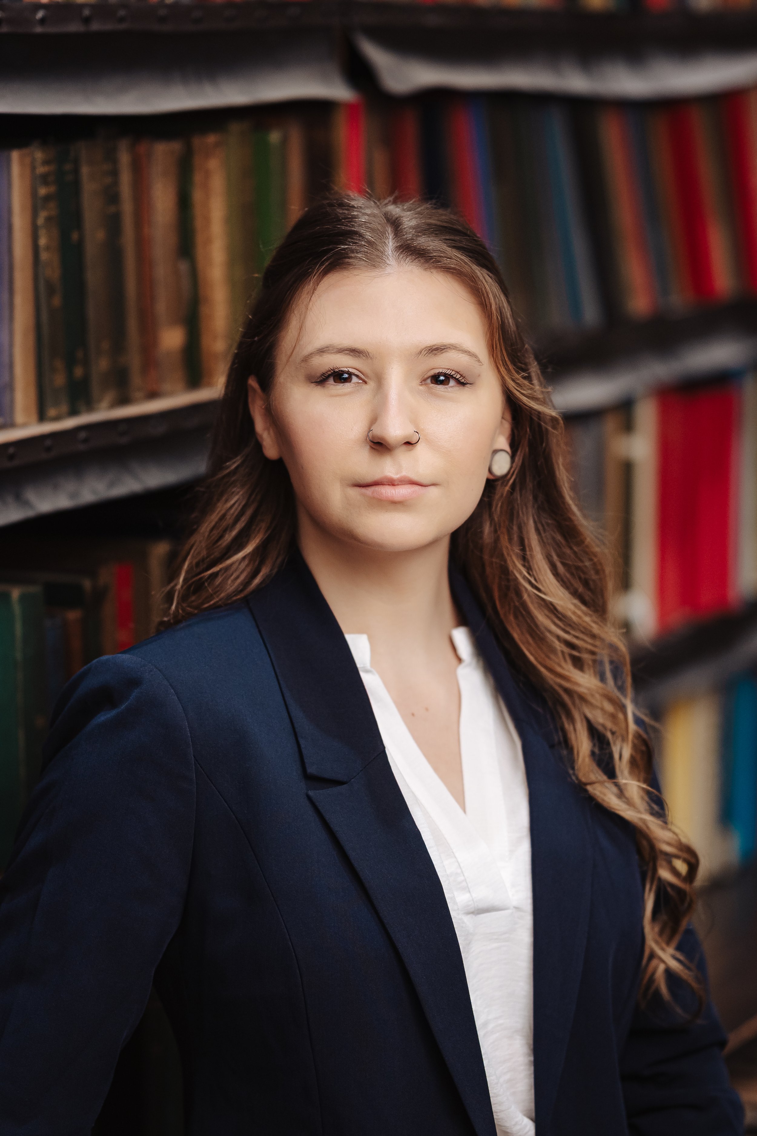 A young woman with wavy brown hair, wearing a navy blazer and white blouse, standing in front of a bookshelf filled with colorful books.