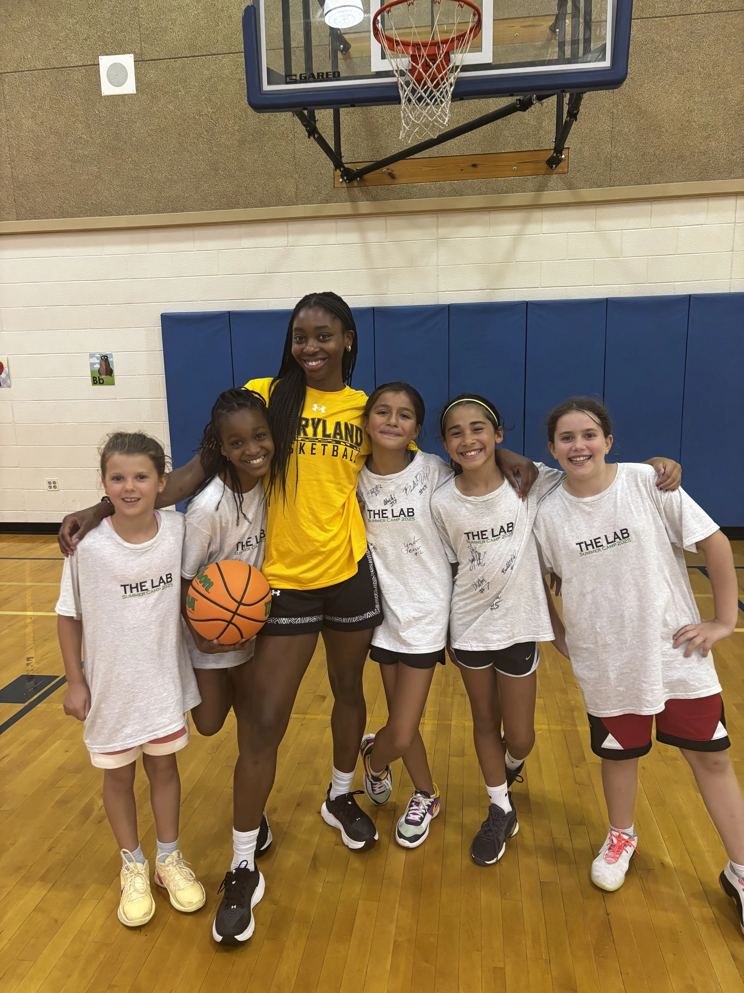 Group of six girls posing on a basketball court with a ball, wearing athletic clothes, with a basketball hoop above them.