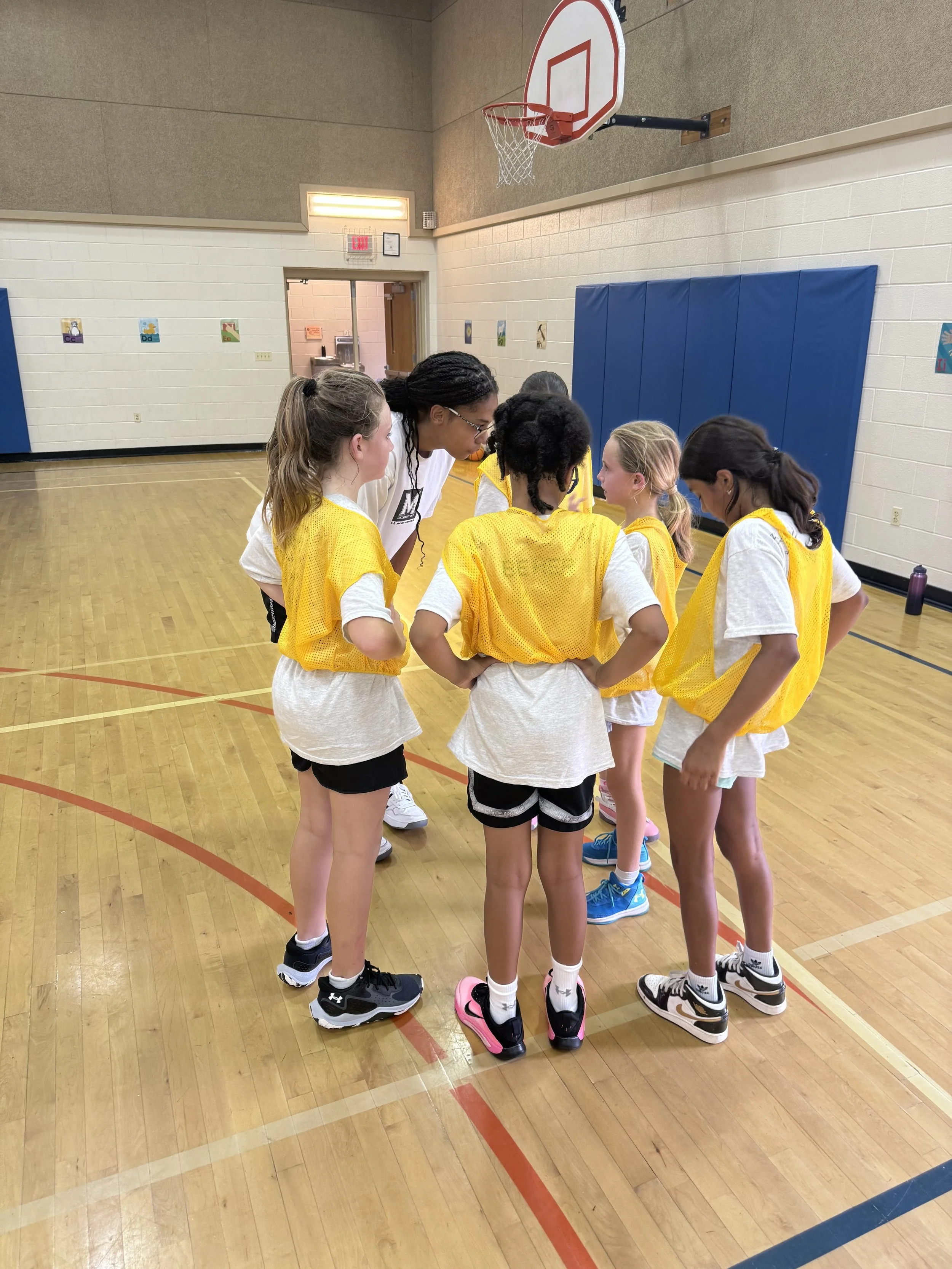 A group of young girls in yellow practice jerseys and white shirts huddle together in a gymnasium, likely discussing strategy during a basketball practice.