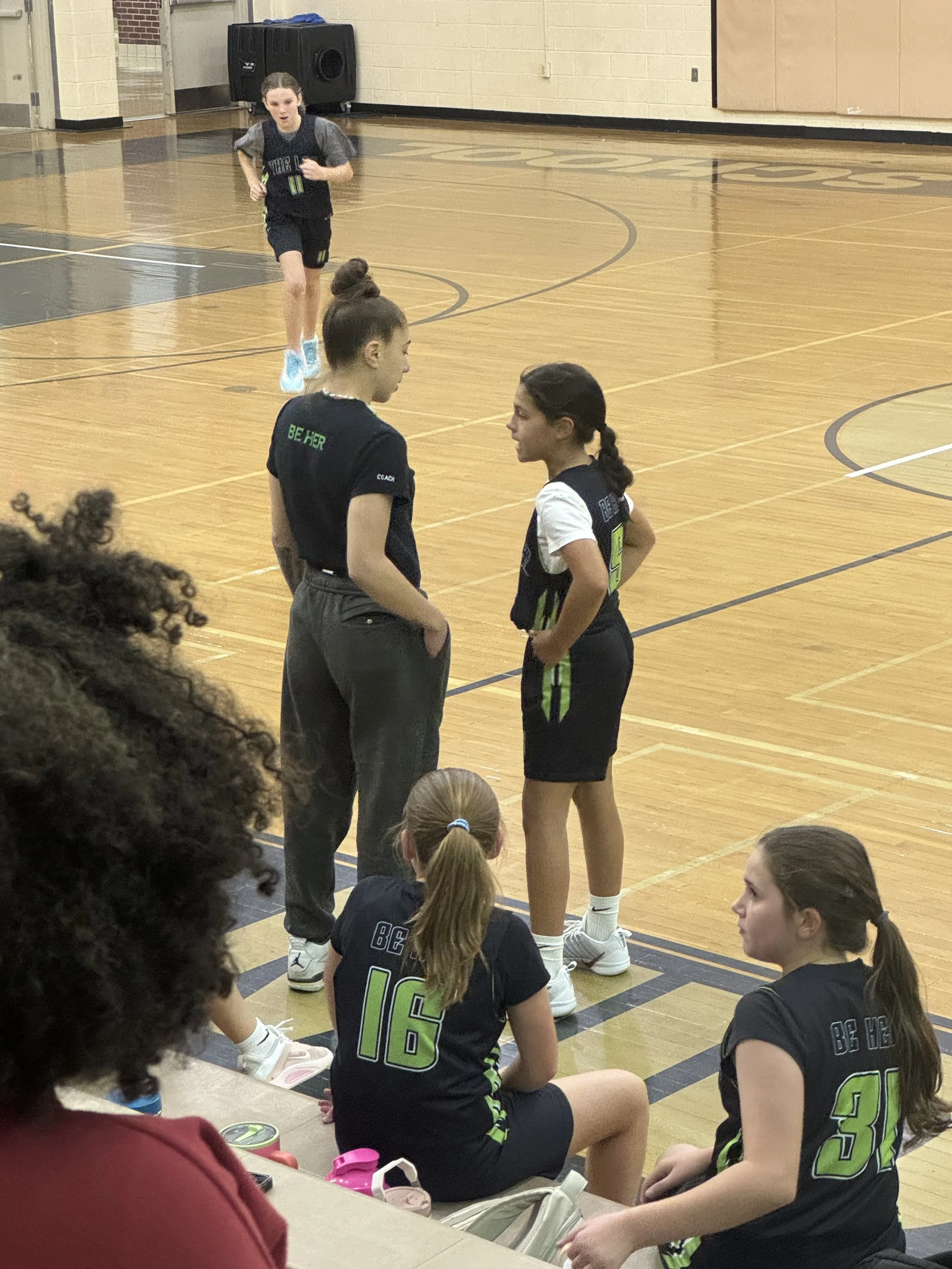 A high school girls basketball team coach talking to players sitting on a gymnasium floor during a break in the game.