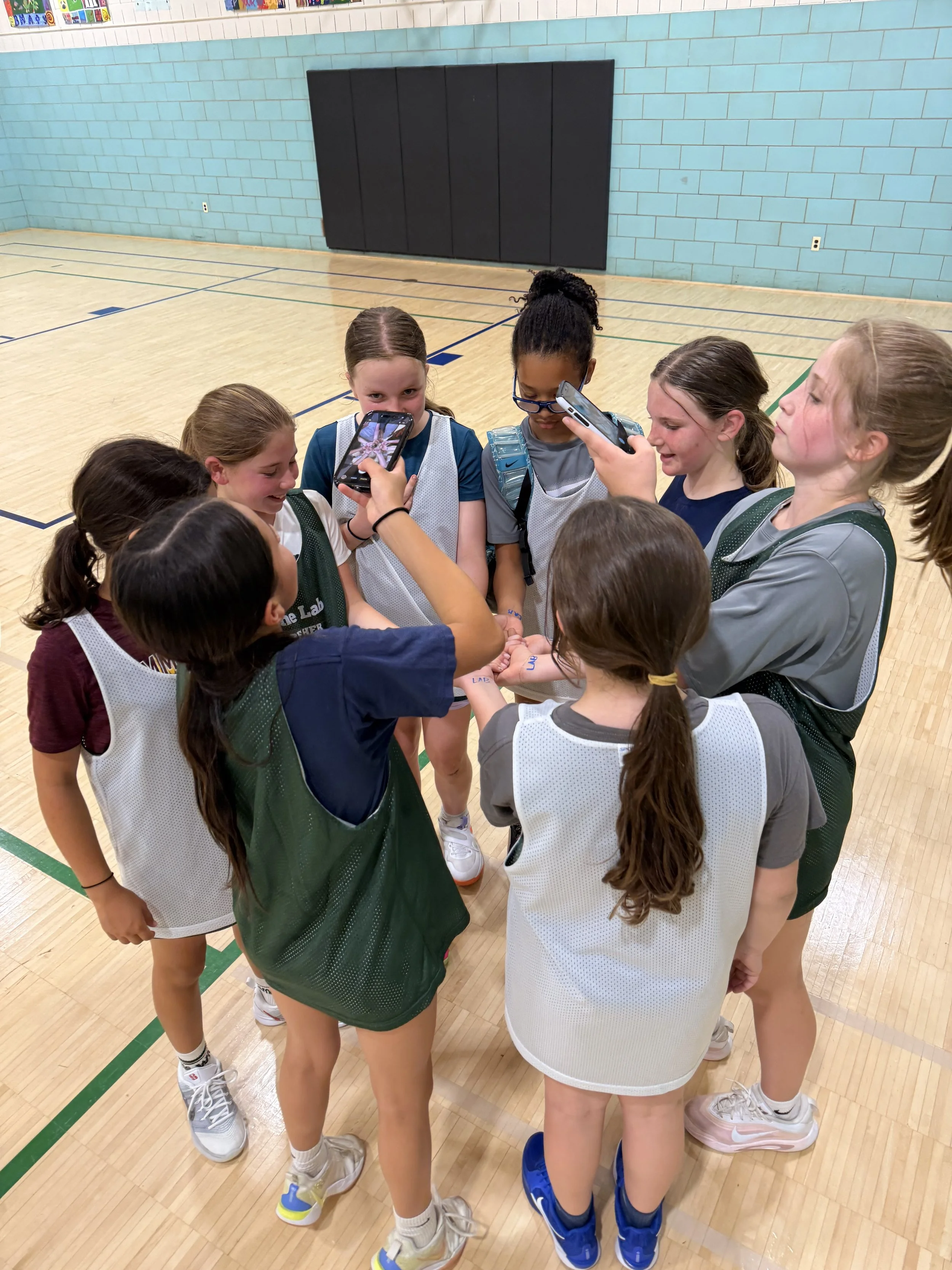 A group of young girls in basketball uniforms huddled together on an indoor basketball court, some holding or taking pictures with smartphones, involving a post-game celebration or team talk.