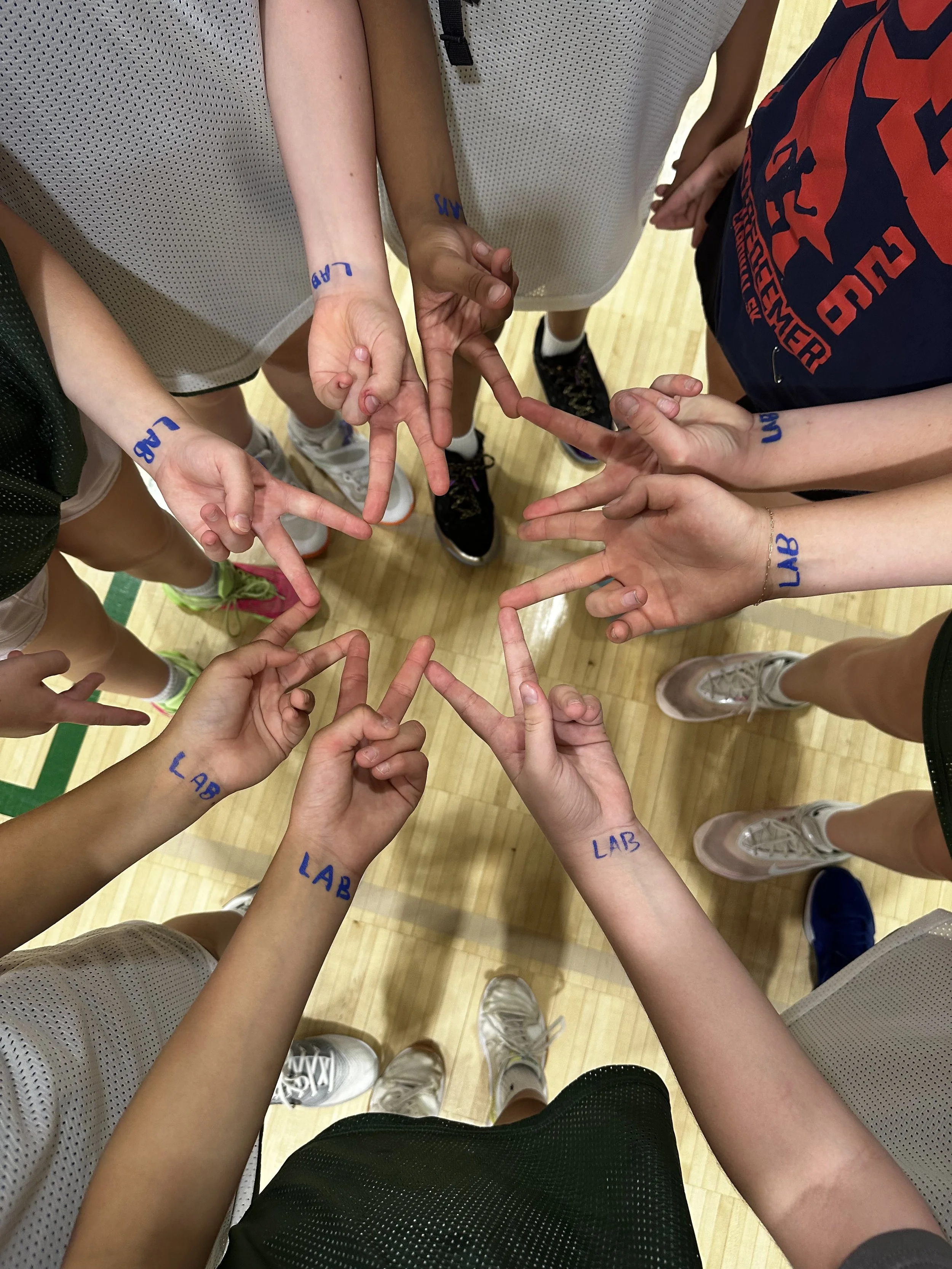 A group of people in a gym wearing basketball uniforms, forming a circle with their hands with the word 'LAB' written on each person's arm.