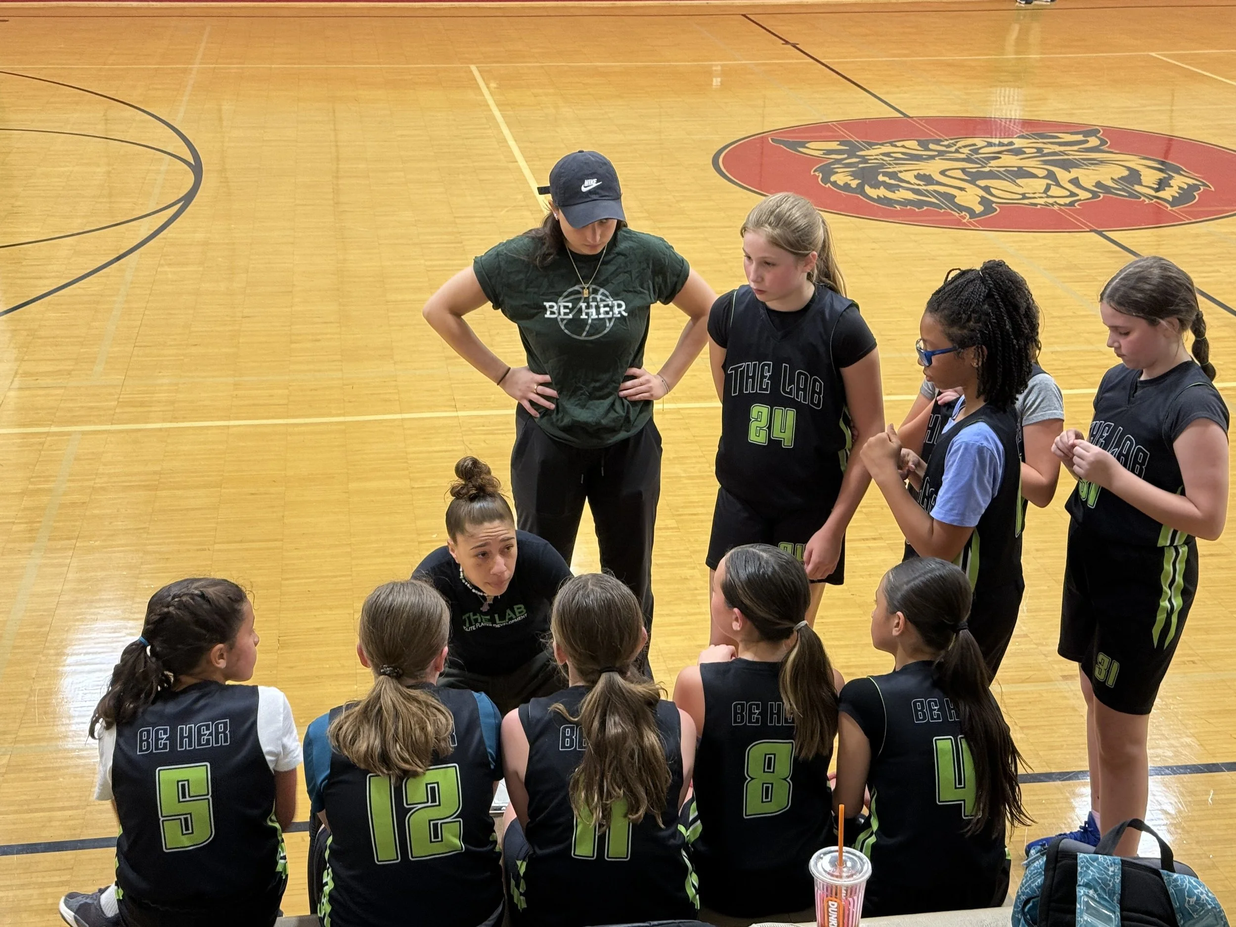 A group of young female basketball players wearing black uniforms with green numbers and the words "Be Her" on the back, sitting and standing on a basketball court, listening to a coach or team leader.