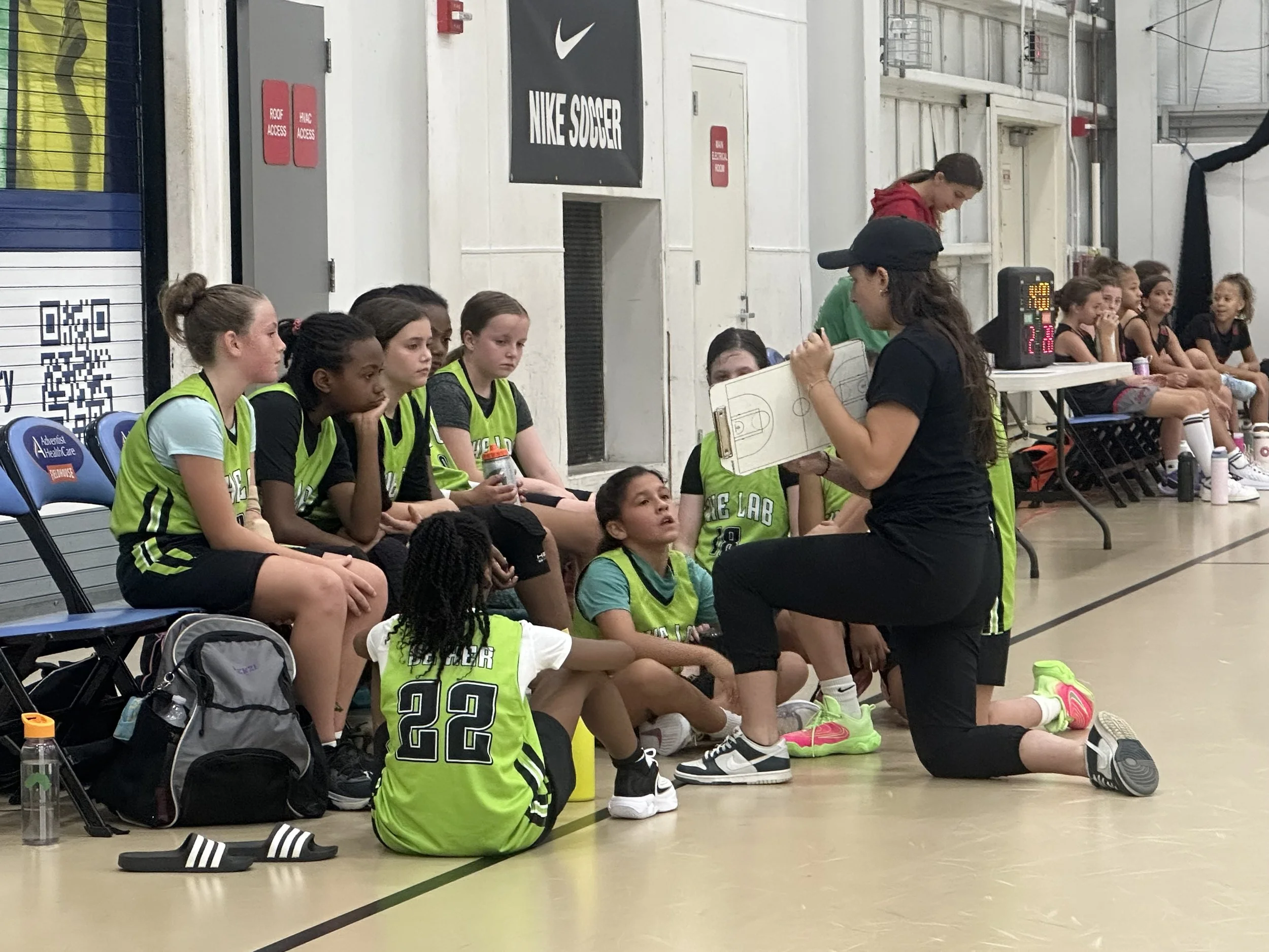 A youth basketball team listens to their coach during a timeout on the sidelines of a game. The players are seated on chairs and the floor, wearing green jerseys, with a coach kneeling and speaking to them. A scoreboard is visible in the background, and other players are sitting on a bench.