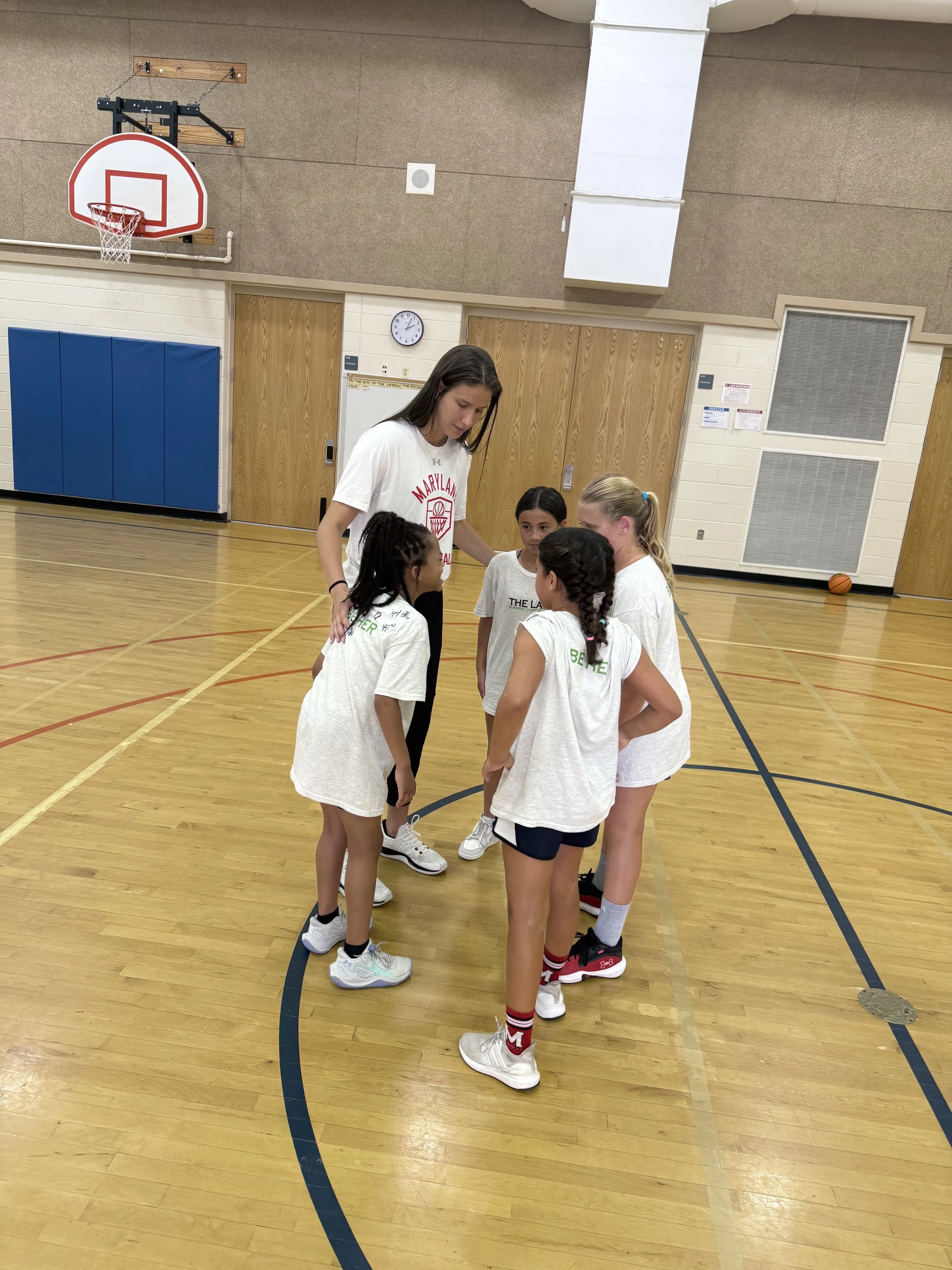 A basketball coach leading a team huddle of young girls on an indoor basketball court.