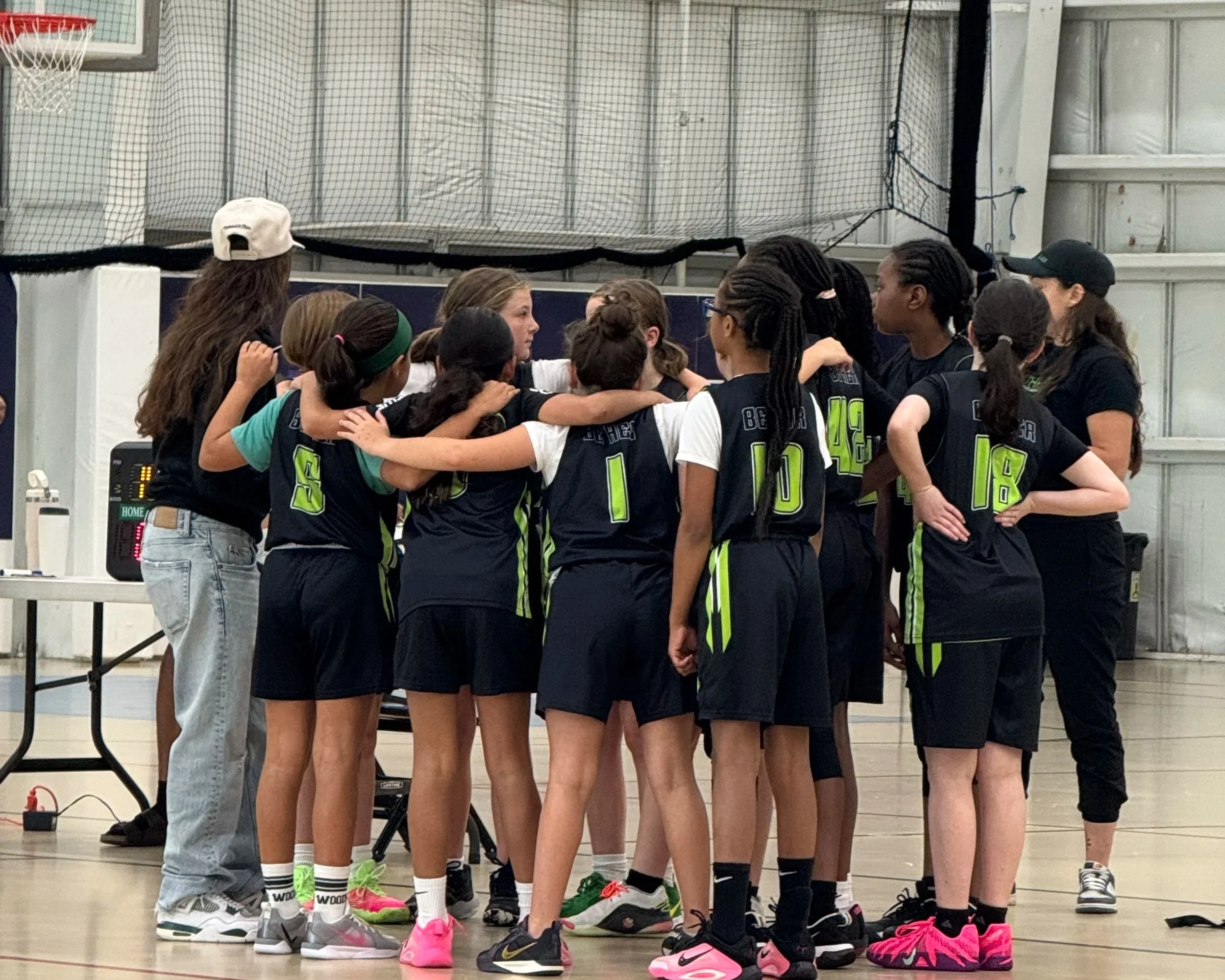 A girls' basketball team in black uniforms with neon green numbers and stripes is huddled together on a court, listening to their coach during a timeout.