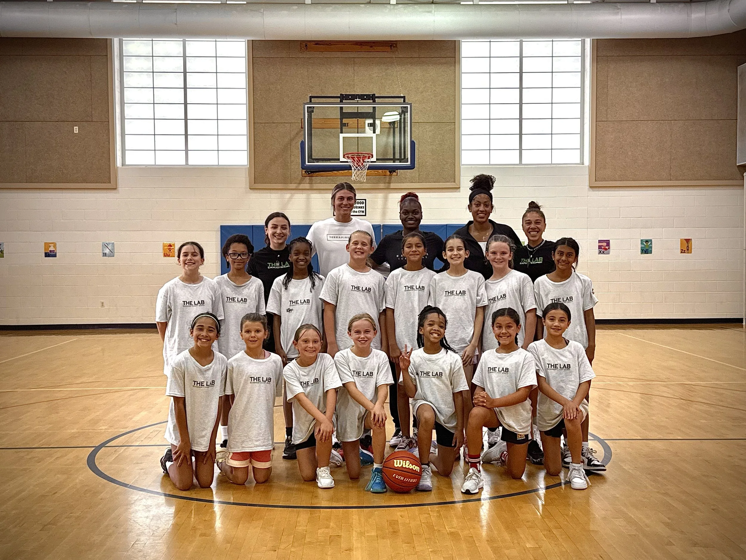 A group of young girls and their coaches or teachers posing for a photo in a gymnasium. The girls are wearing matching white T-shirts with "THE LAB" printed on them, and there is a basketball on the floor in front of them.