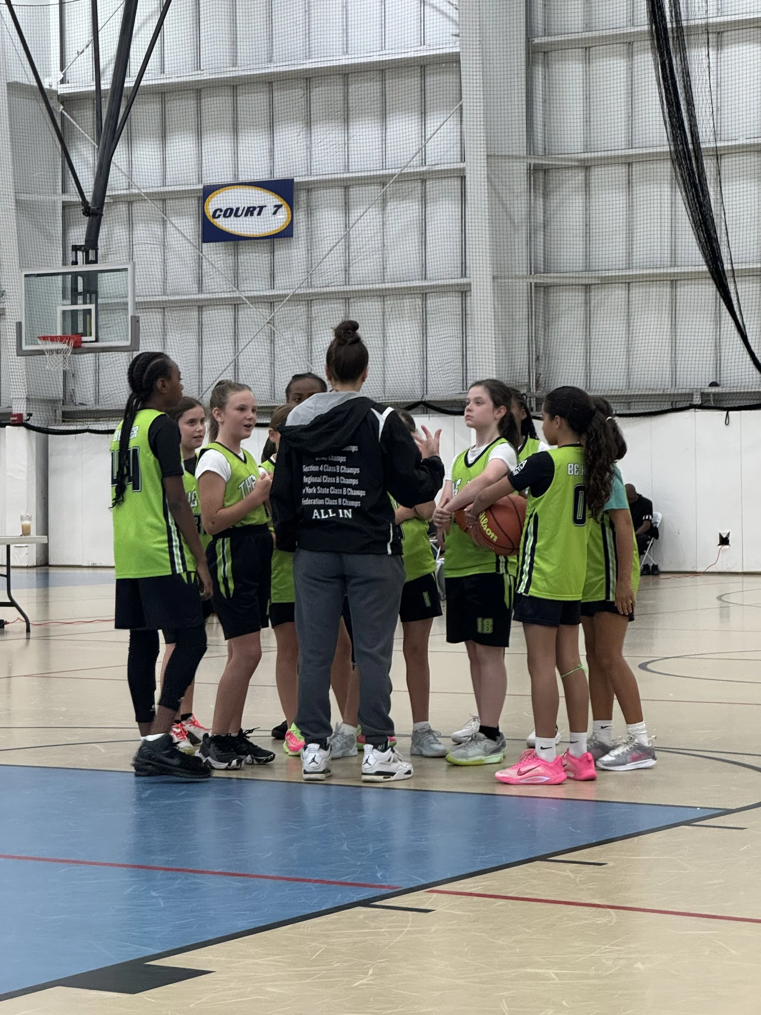 Girls' youth basketball team gathered around their coach in a gymnasium during a timeout.
