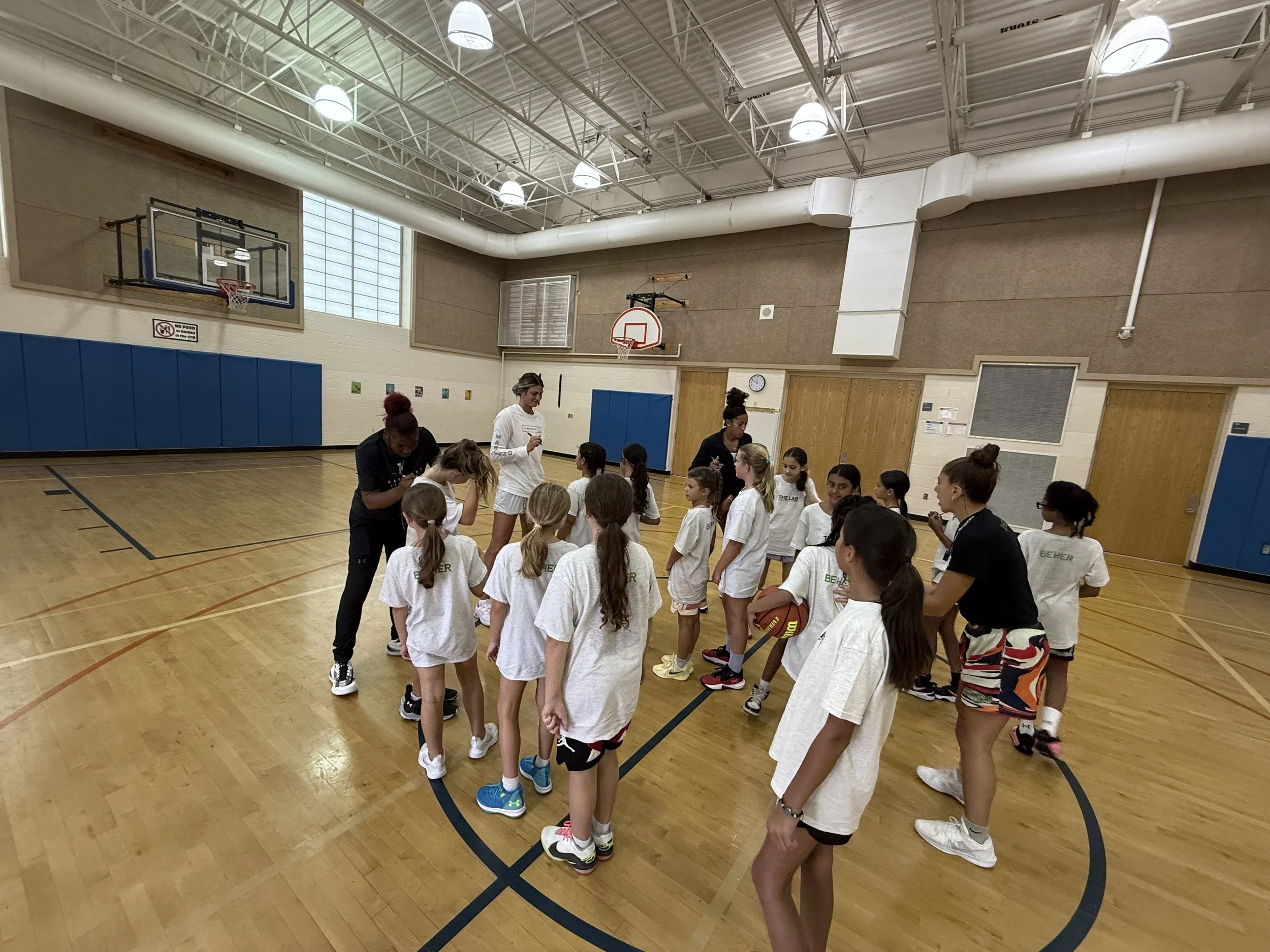A girls' basketball team in a gymnasium during a practice or game, with two coaches giving instructions.