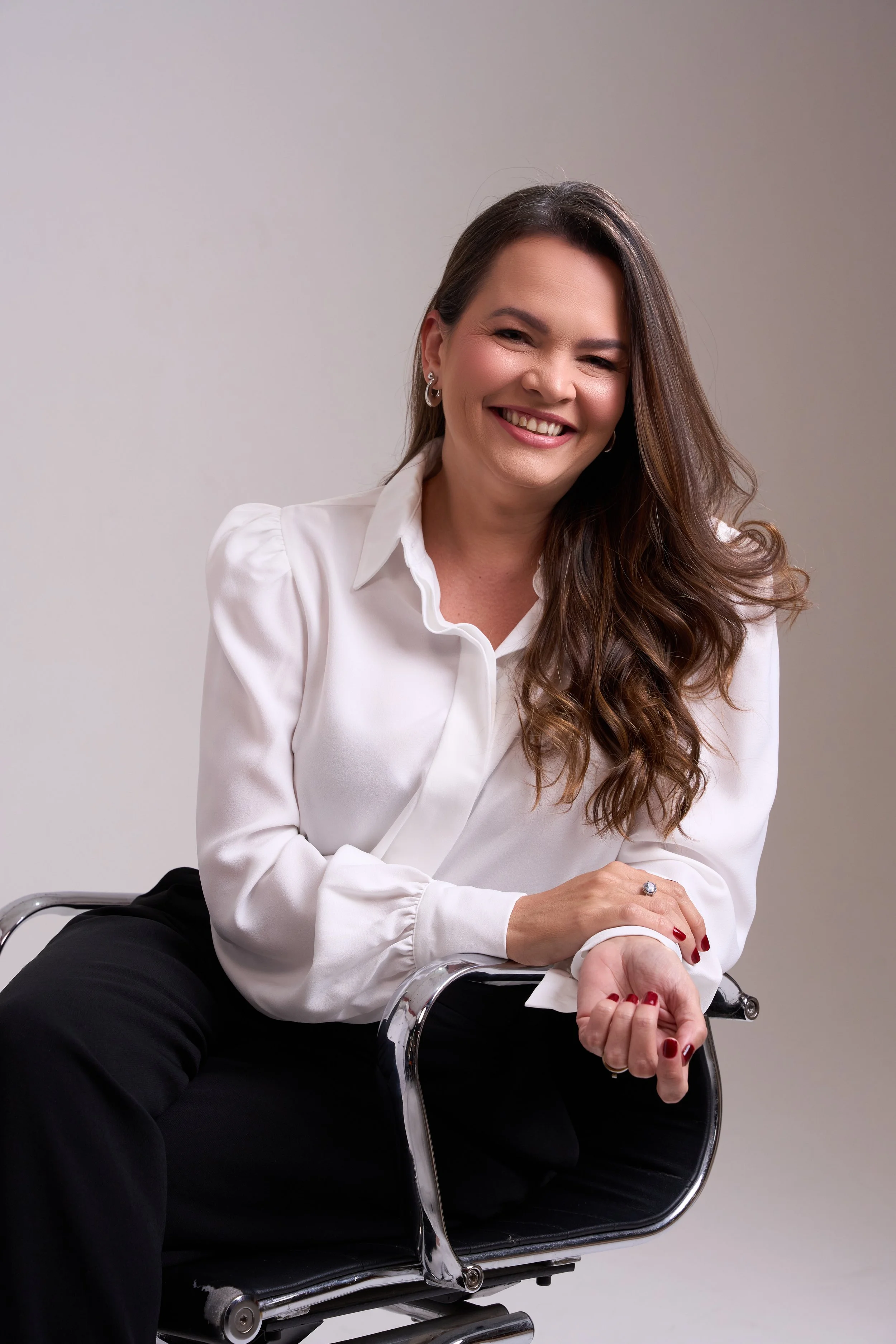 Mulher sorrindo, com cabelo castanho, usando camisa branca e calças pretas, sentada em cadeira de escritório, em fundo neutro.