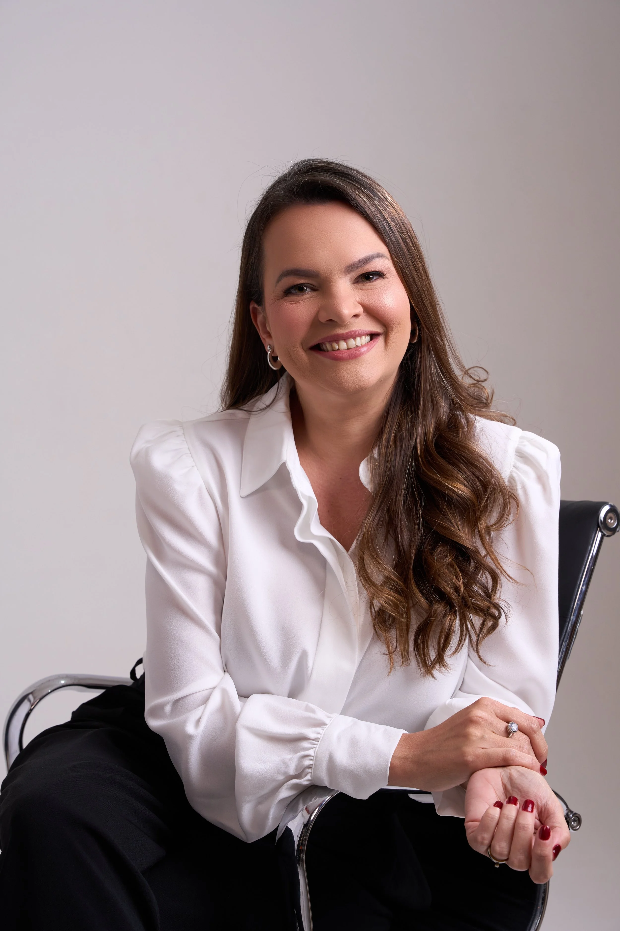 Mulher sorridente com cabelo castanho, usa blusa branca de manga bufante, sentada em cadeira de escritório de metal.