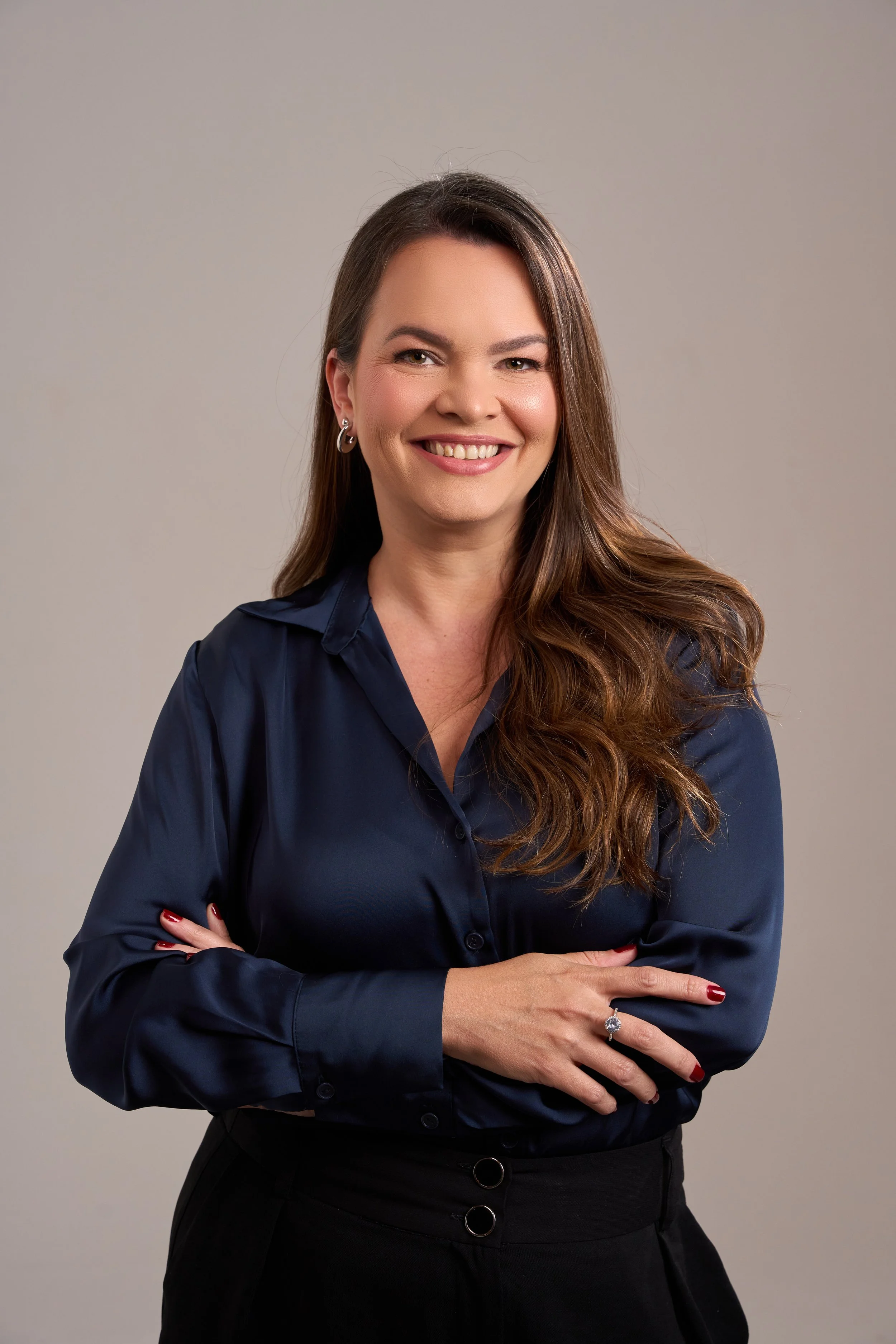 Mulher com cabelos castanhos, sorriso, usando camisa azul escuro, com as mãos cruzadas na frente, de fundo neutro.