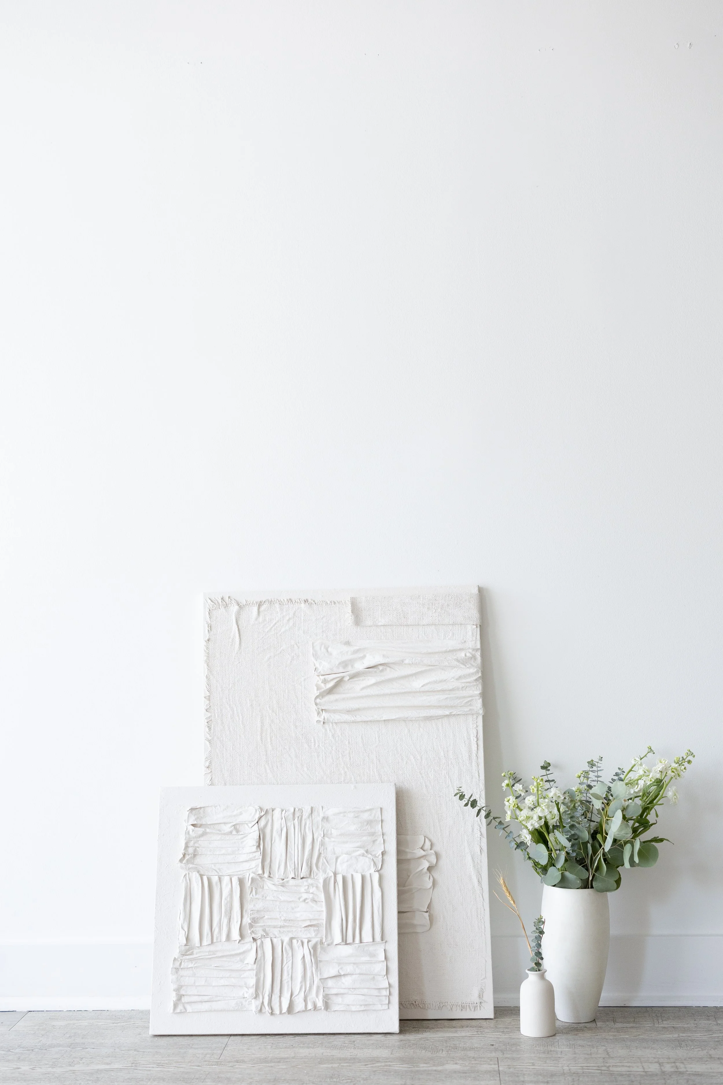 Two textured white art canvases leaning against a white wall, one smaller and one larger, next to a white vase with greenery and small white flowers.