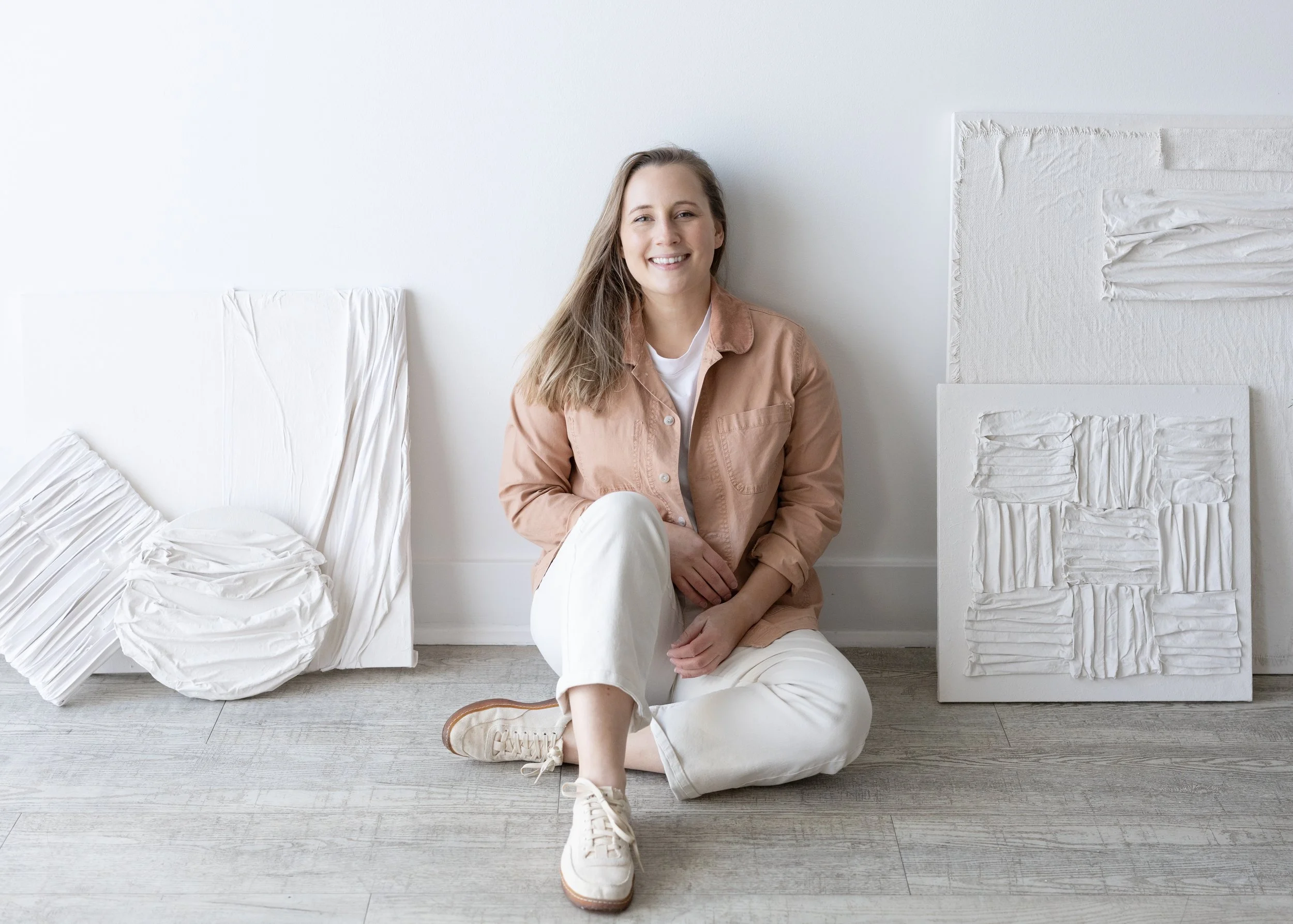 A woman sitting on the floor with white and beige textured art pieces around her, smiling at the camera.