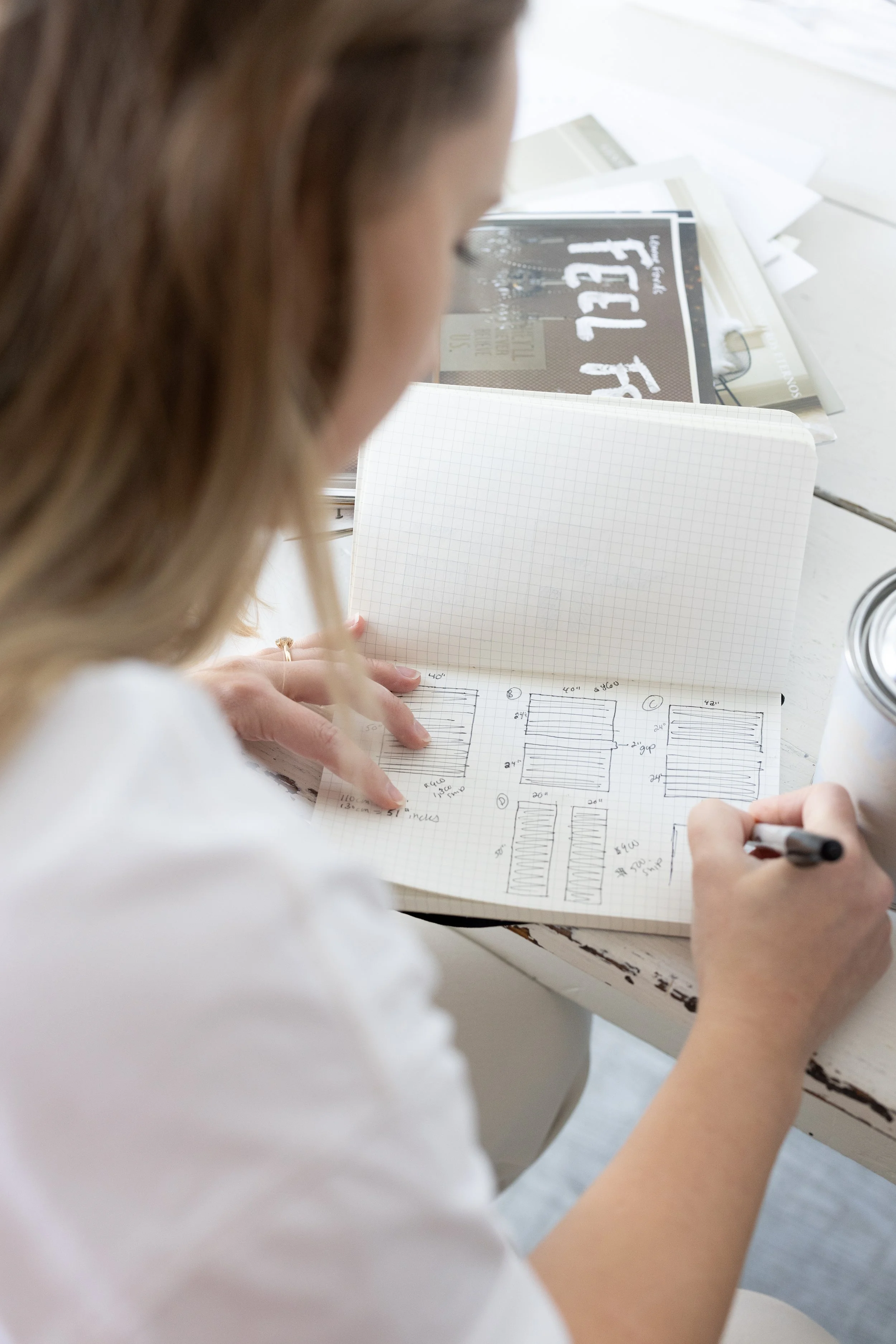 A woman with light brown hair sitting at a white table, sketching and taking notes in a graph paper notebook, with various papers and a can on the table.