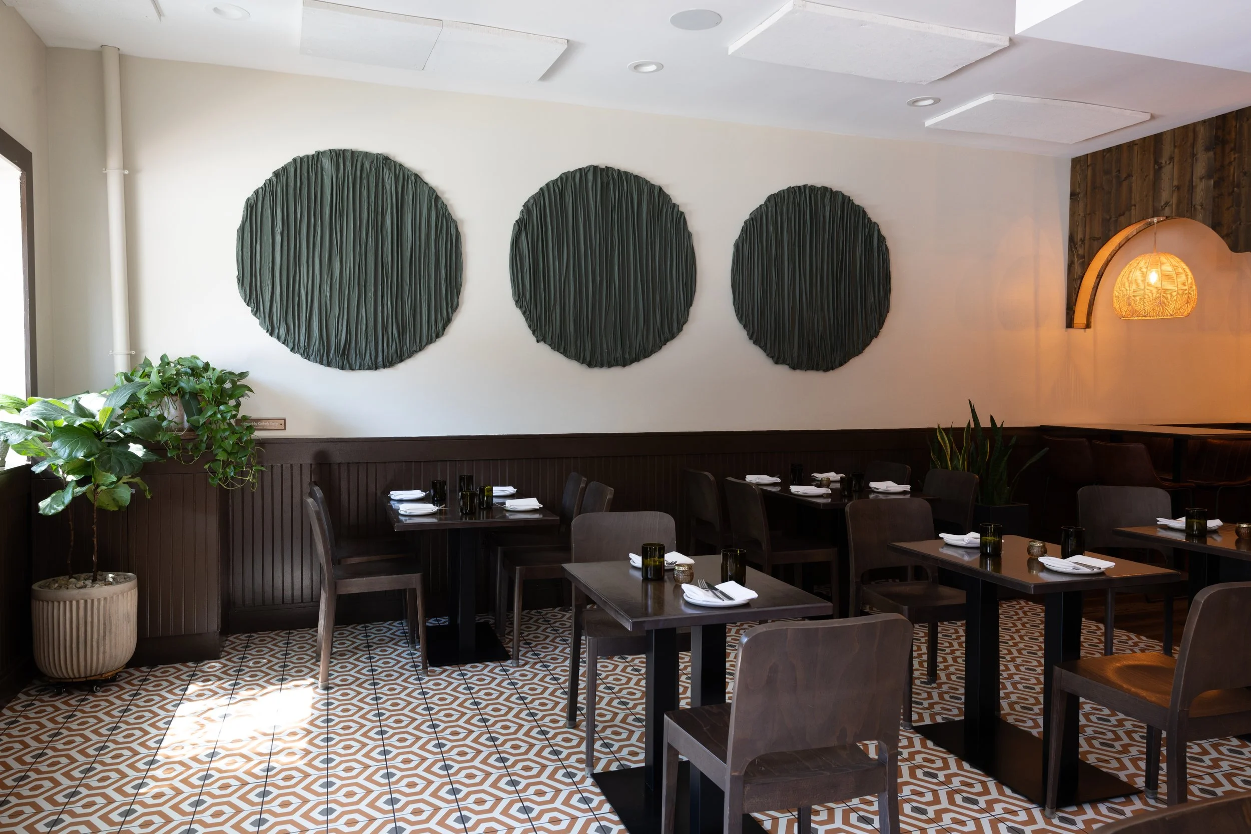 Empty restaurant dining area with black tables, chairs, and green wall art, featuring a potted plant near the window and patterned floor tiles.