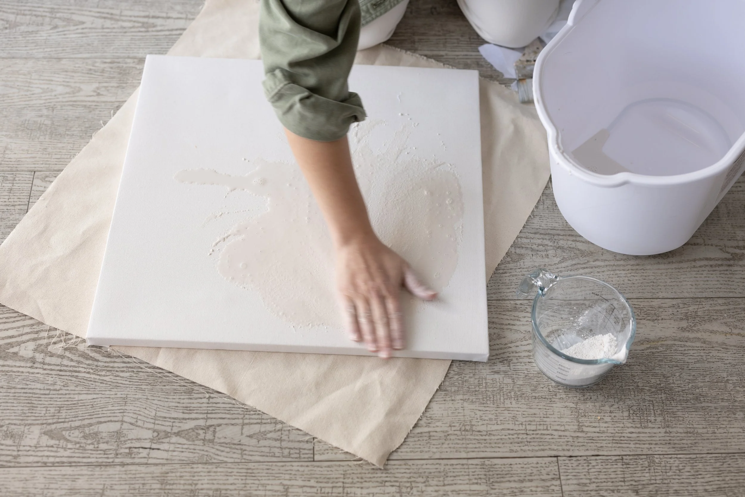 Person working on a canvas with water, a measuring cup of white powder, and an empty water container nearby.