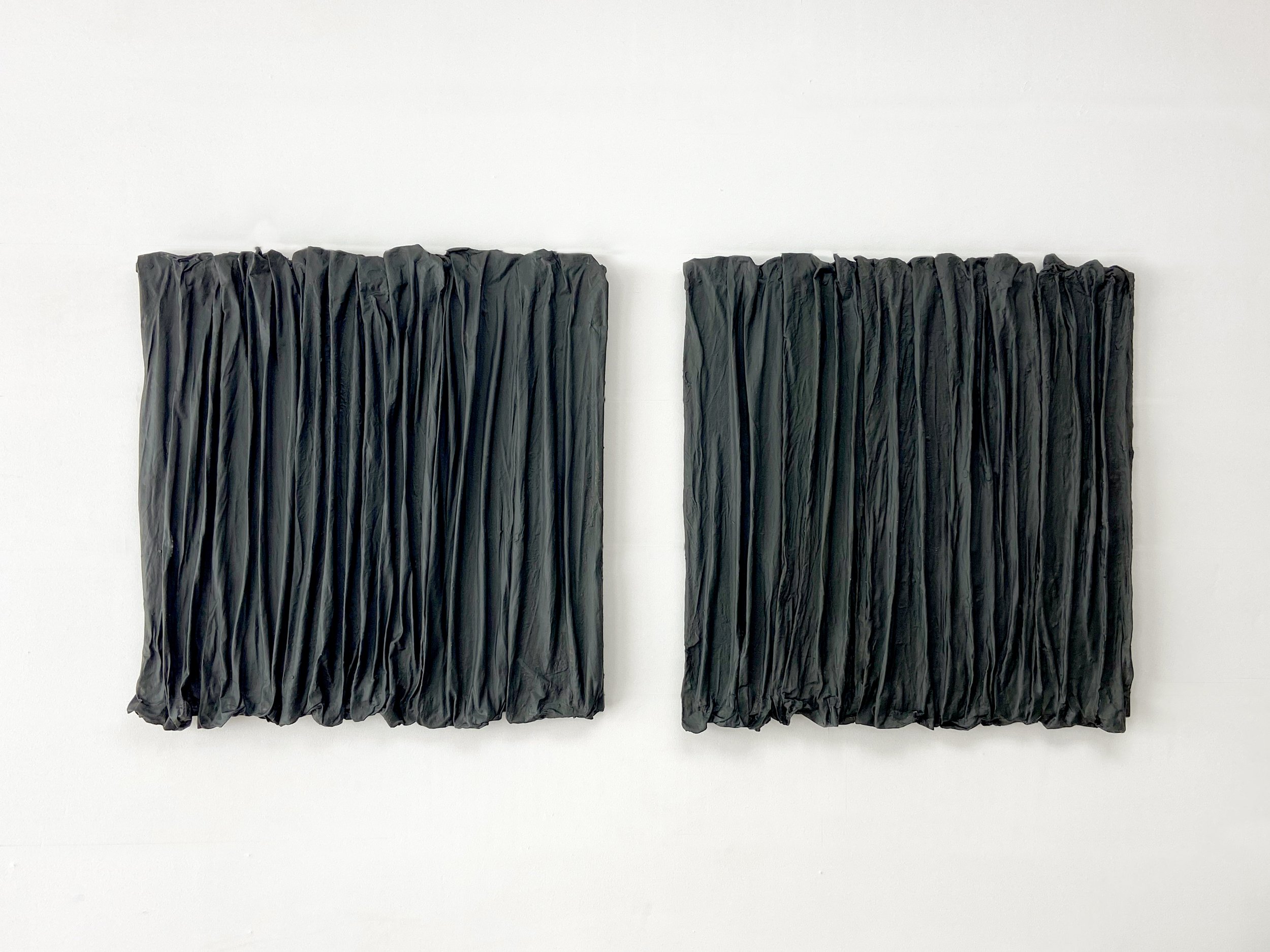 Two square black textured artworks with a crinkled appearance hung on a white wall.