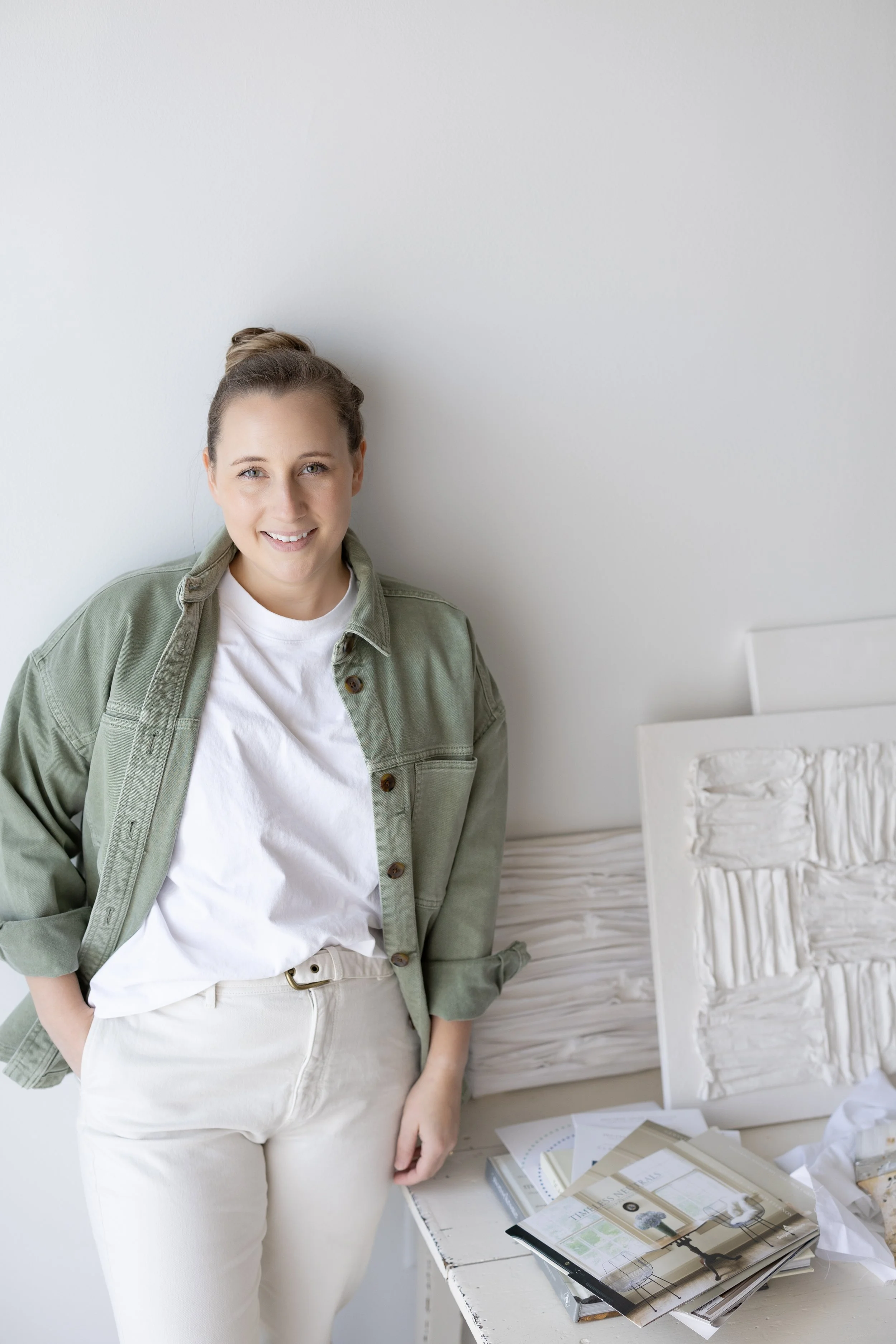 A smiling woman standing in a bright, minimalist room with a white wall background, wearing a white t-shirt, beige pants, and an olive green jacket, with artwork and papers on a table beside her.