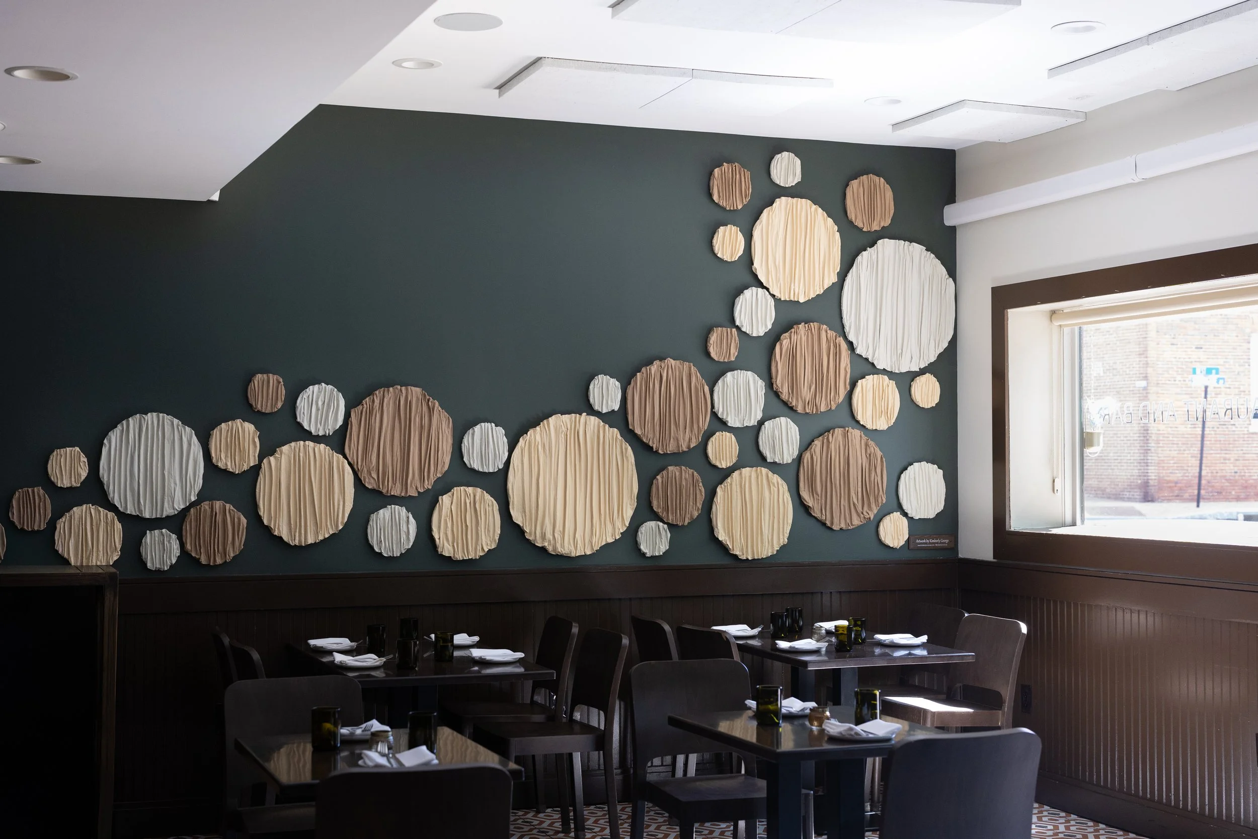 Dining room with dark tables and chairs, wall decor of large and small round textured fabric art pieces in neutral tones, and a large window letting in natural light.