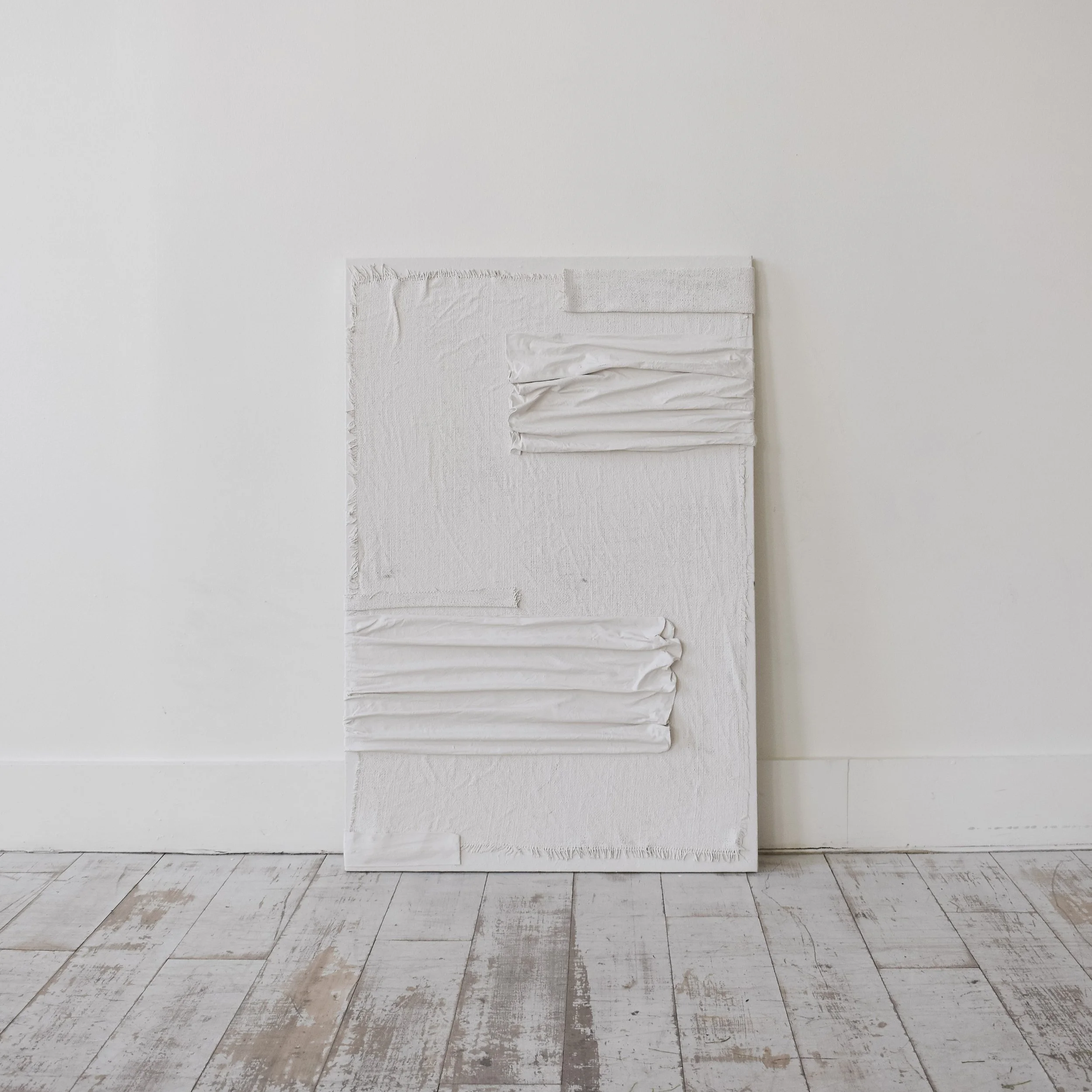 Abstract white textured art piece with layered fabric and paper elements, leaning against a white wall on a whitewashed wooden floor.