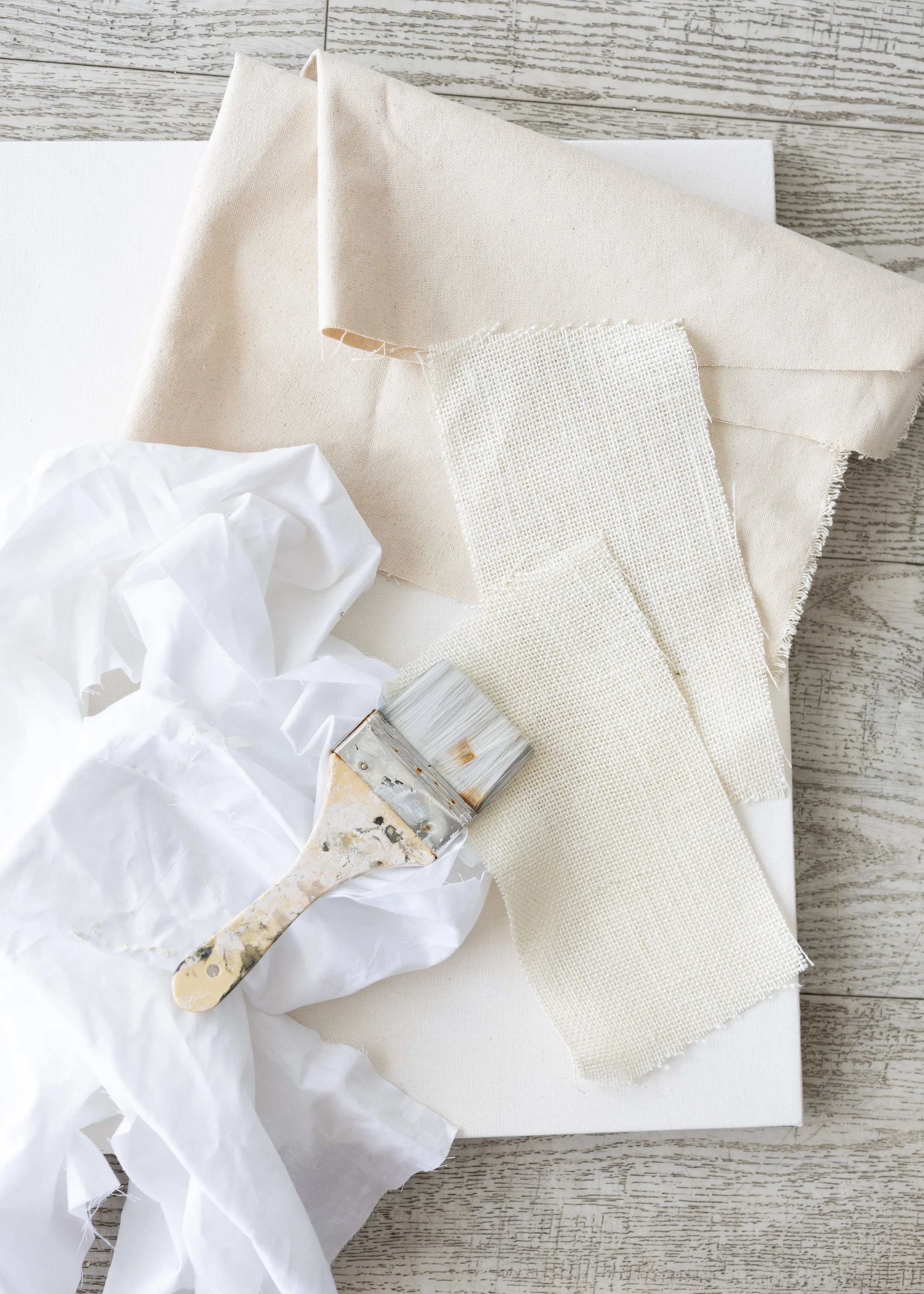 An assortment of fabric swatches, a paintbrush, and tissue paper on a wooden surface, suggesting a craft or painting project.