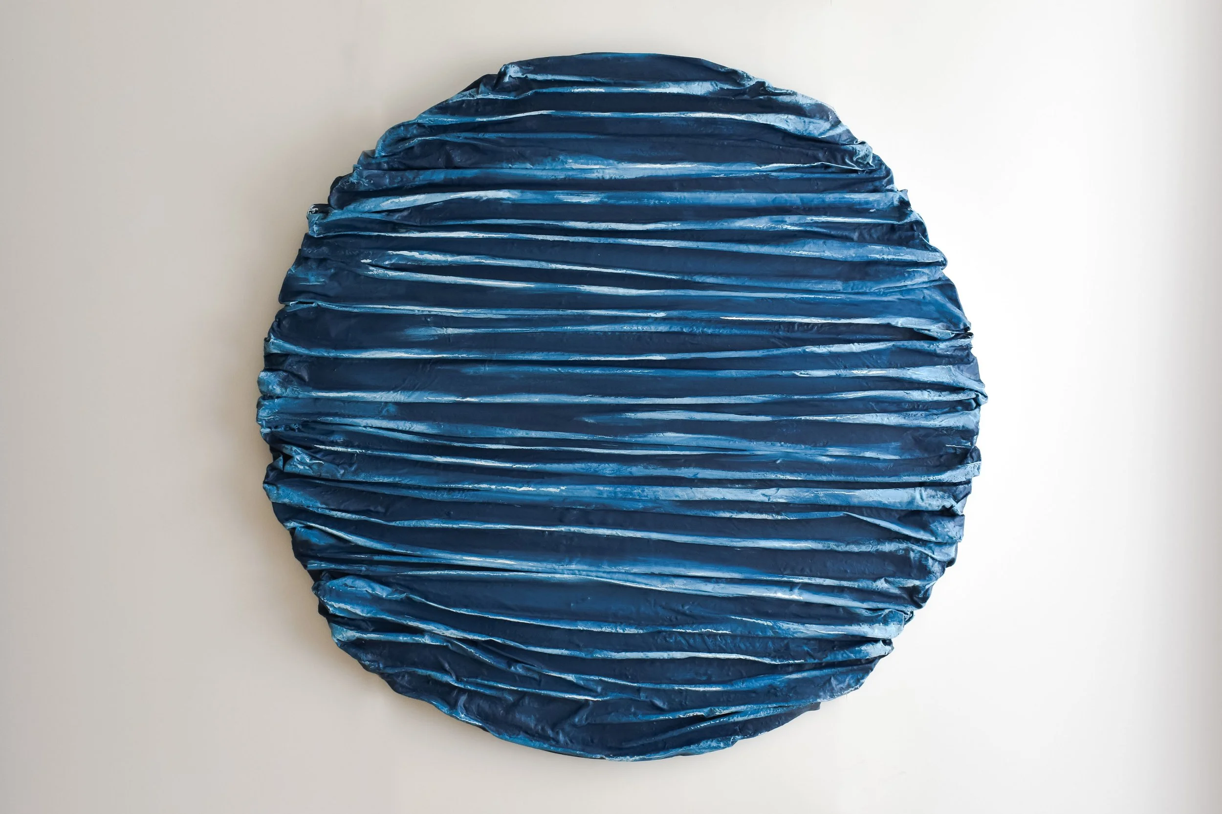 Blue, round, textured sculpture with horizontal ridges on a white background.
