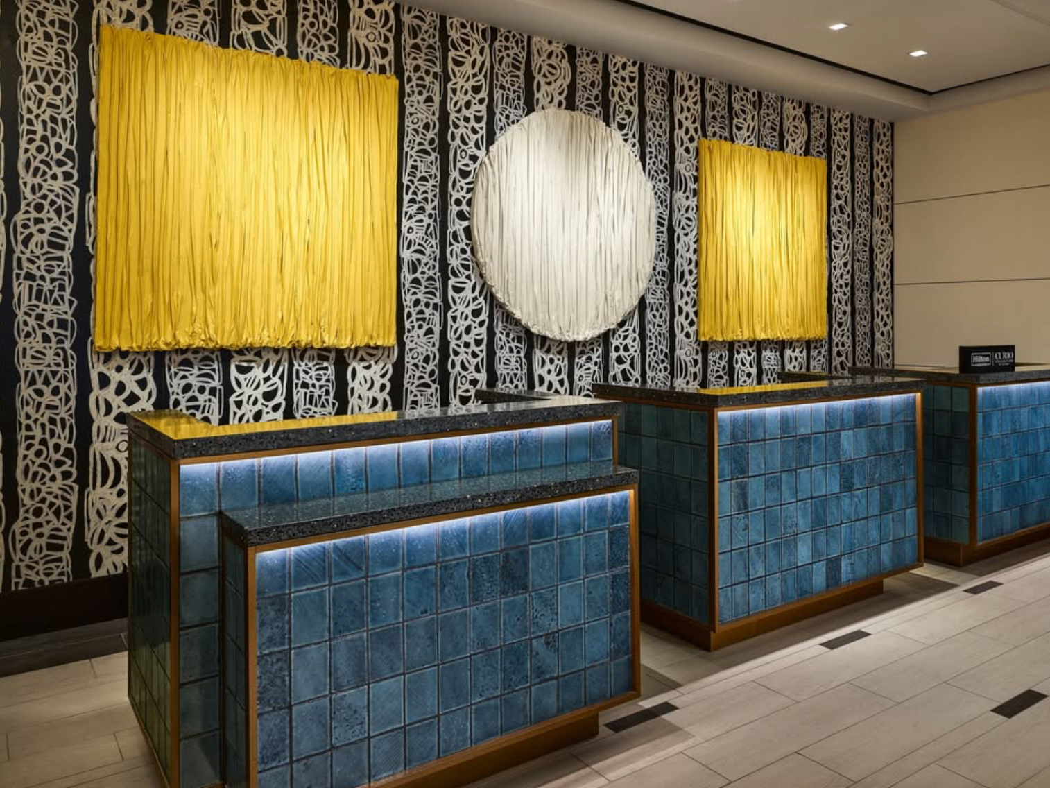 Hotel or reception area with three blue tiled counters with black countertops, yellow fabric wall hangings, and black and white decorative wallpaper.