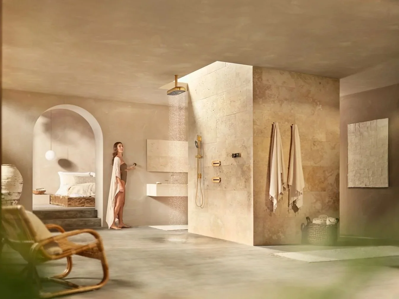 Woman standing in a modern bathroom with a shower, towels, and a bedroom visible through an archway.