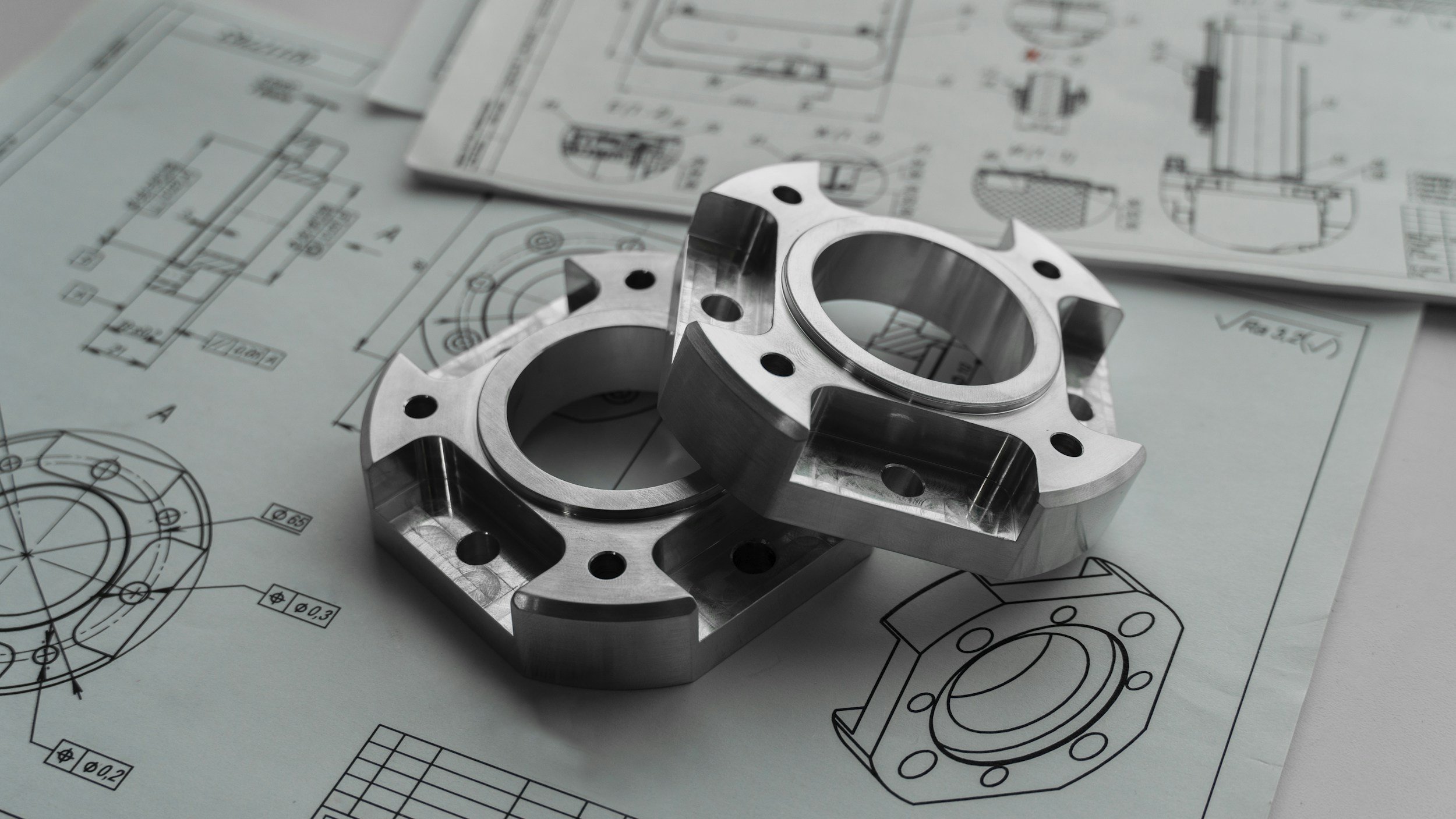 Two machined metal parts placed on engineering blueprints and technical drawings.
