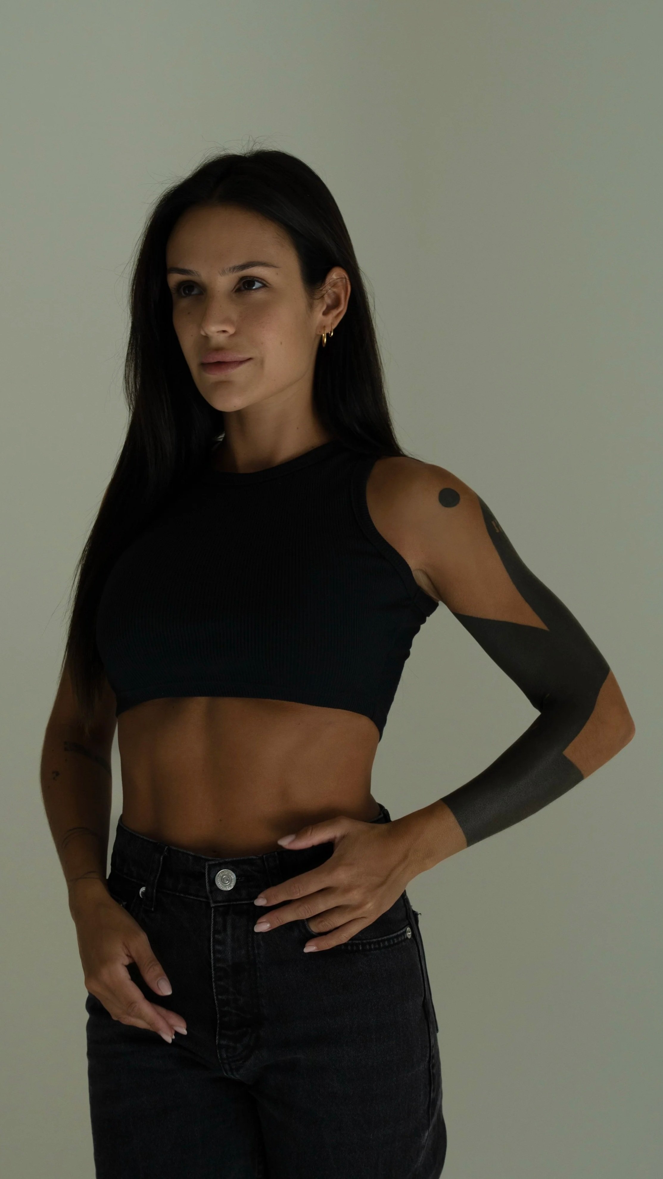 A woman with long dark hair wearing a black crop top, black high-waisted jeans, and black arm tattoo, standing against a plain light-colored wall.