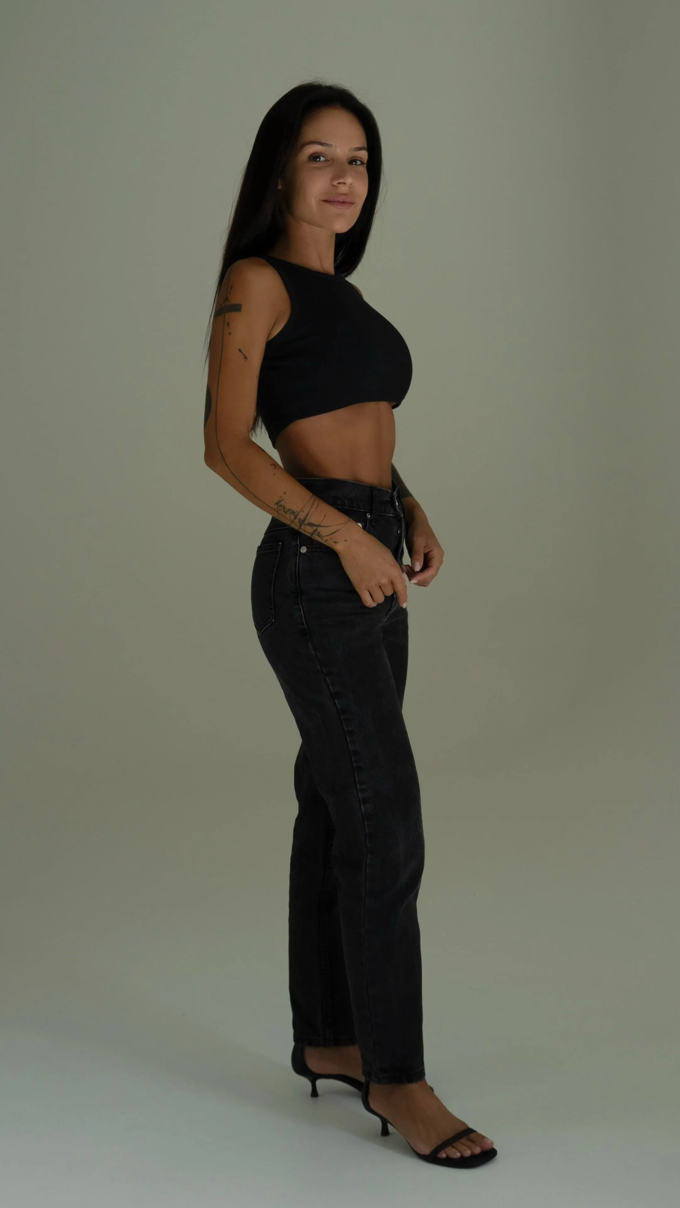 A woman with long dark hair wearing a black sleeveless crop top, black jeans, and black high-heeled sandals, standing against a plain background.