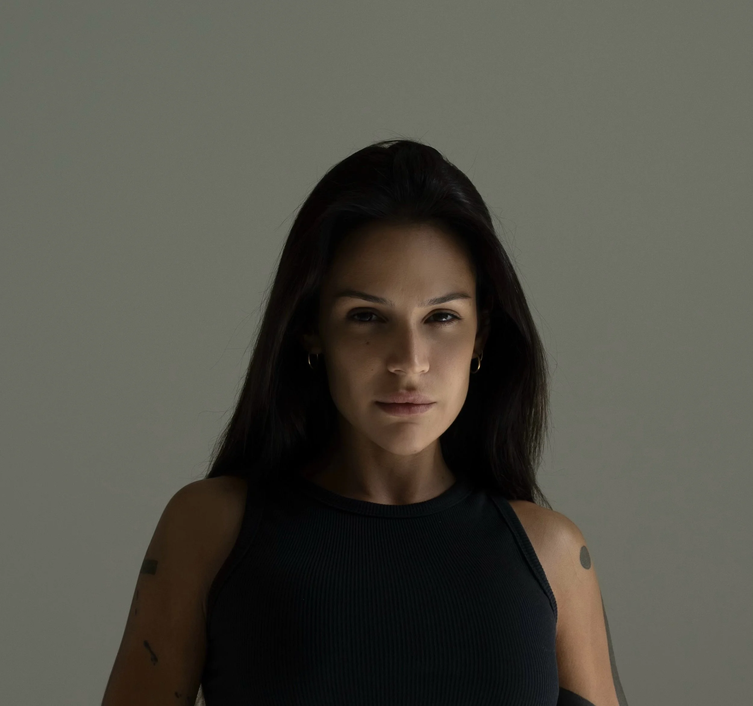 Portrait of a young woman with dark hair, wearing a black sleeveless top, looking directly at the camera with a subtle expression, against a plain gray background.