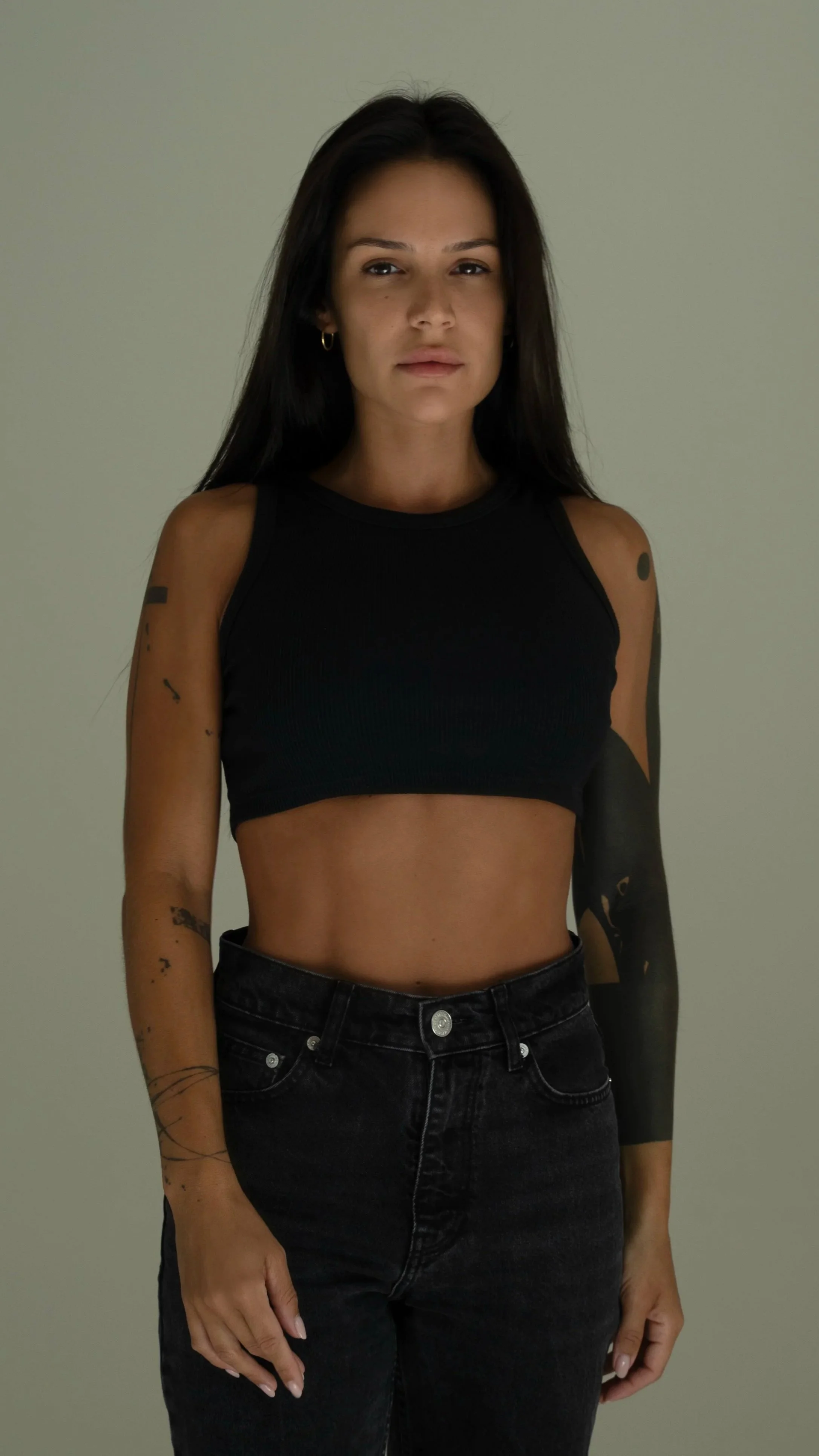 A woman with long dark hair wearing a black crop top and black jeans standing against a plain background.