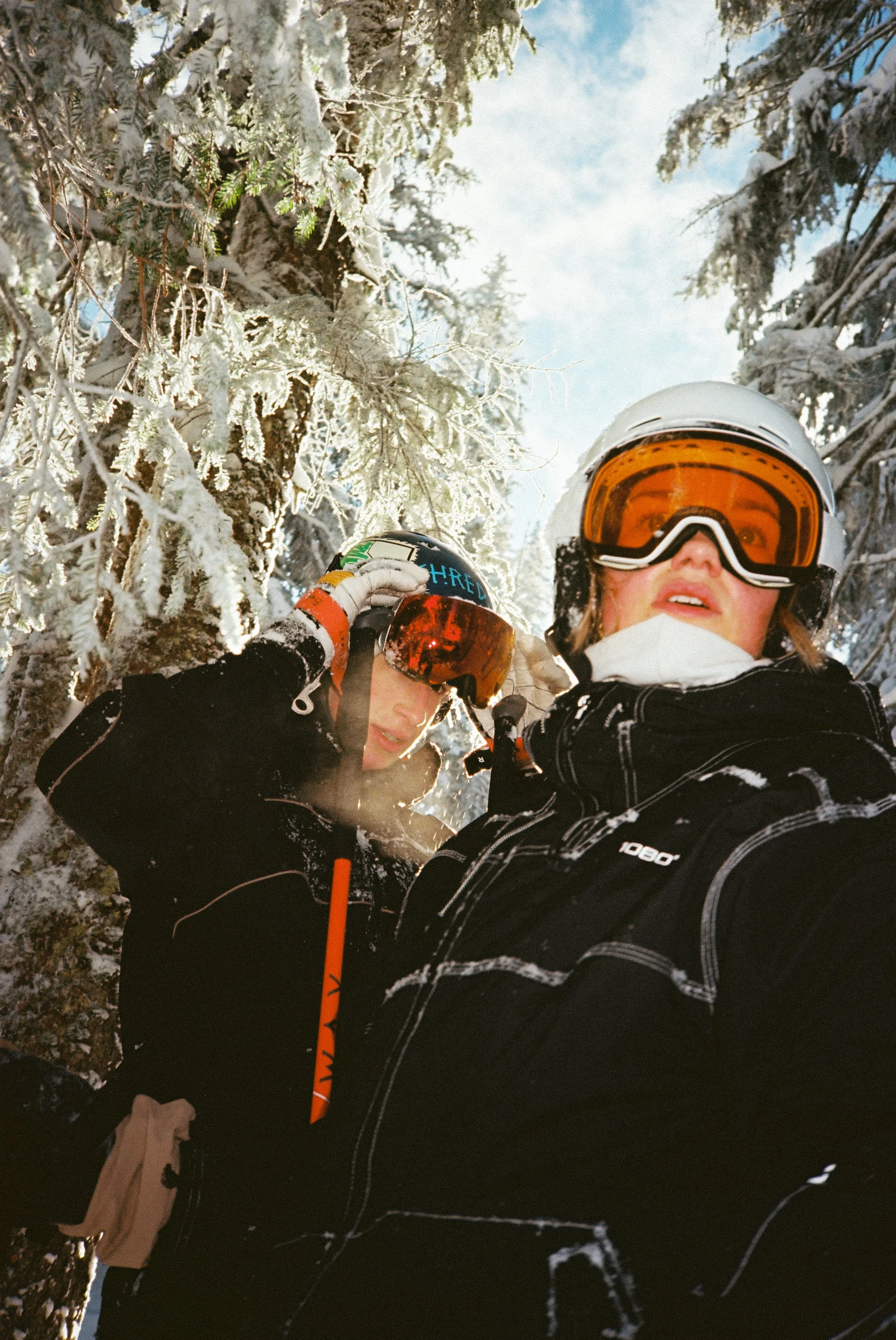 Zwei Skifahrer in Winterkleidung mit Helmen und Skibrillen in verschneitem Wald auf Schneespuren im Schnee.