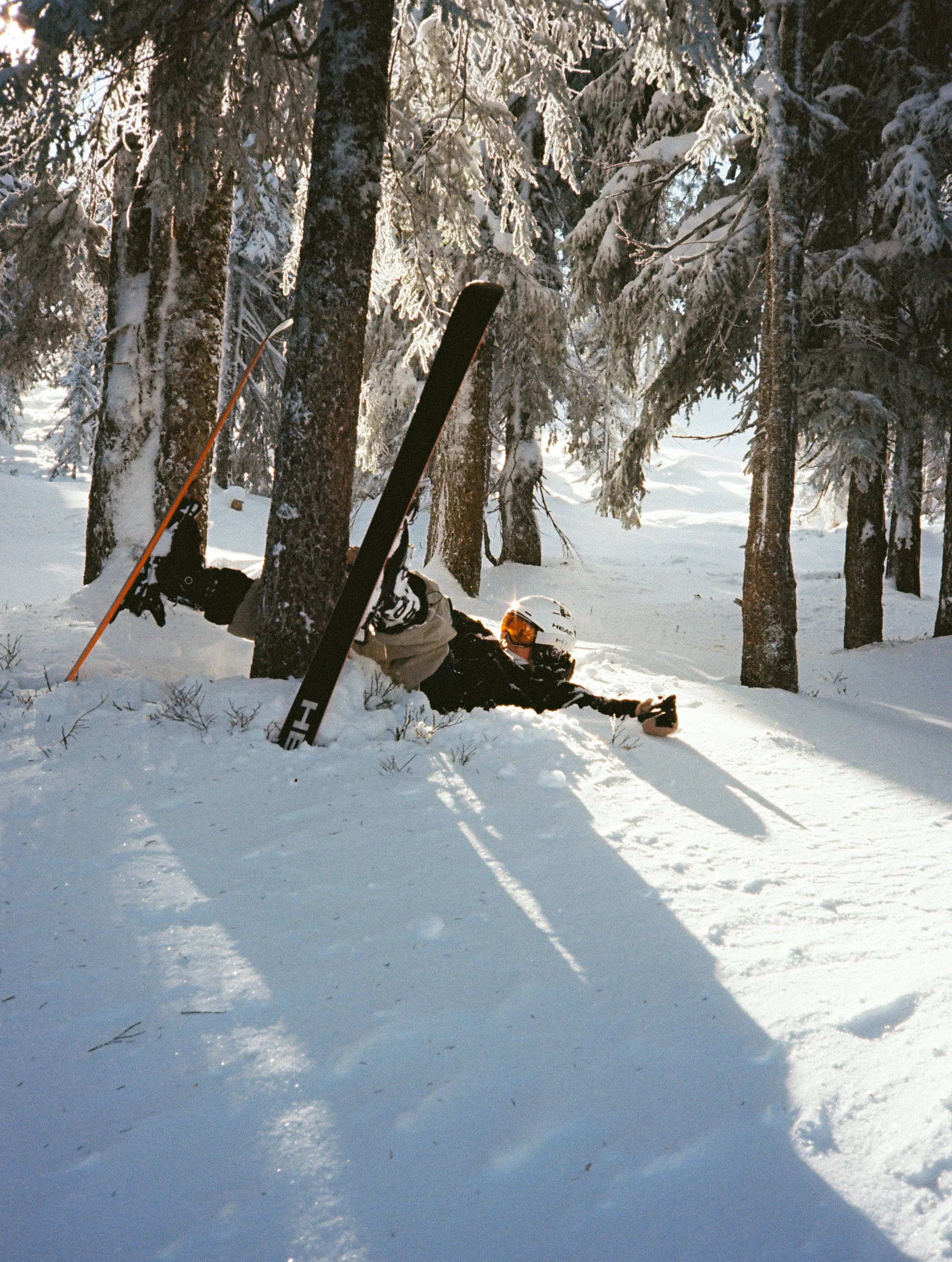 Ein Skifahrer liegt im Schnee im Wald, mit Skiern und Skistöcken, umgeben von Bäumen. Es ist winterlich mit viel Schnee und Sonnenlicht.